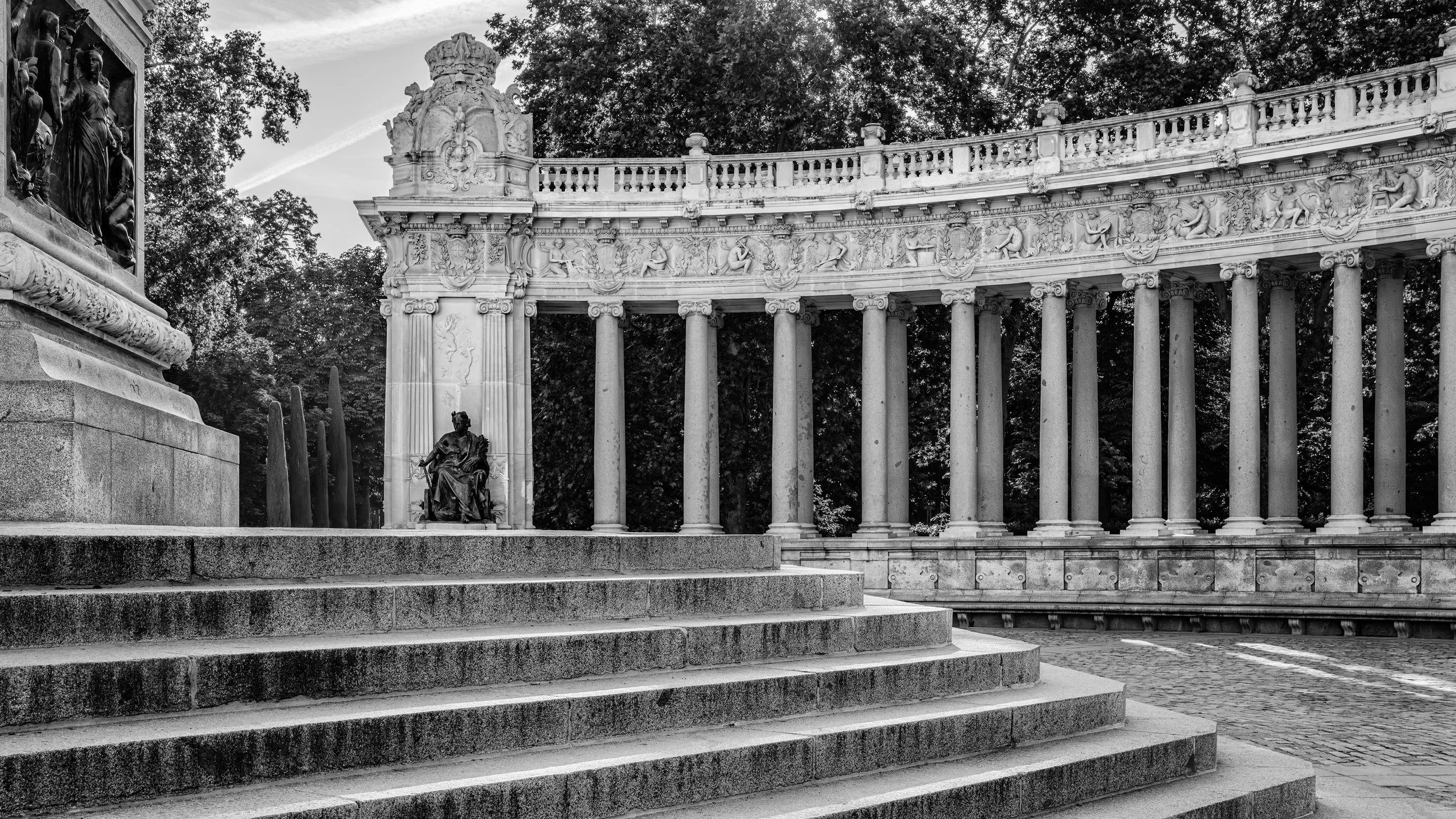 Black and white photograph of a historical monument with steps leading up to a circular colonnade with ornate sculptures and bas-reliefs, surrounded by trees.