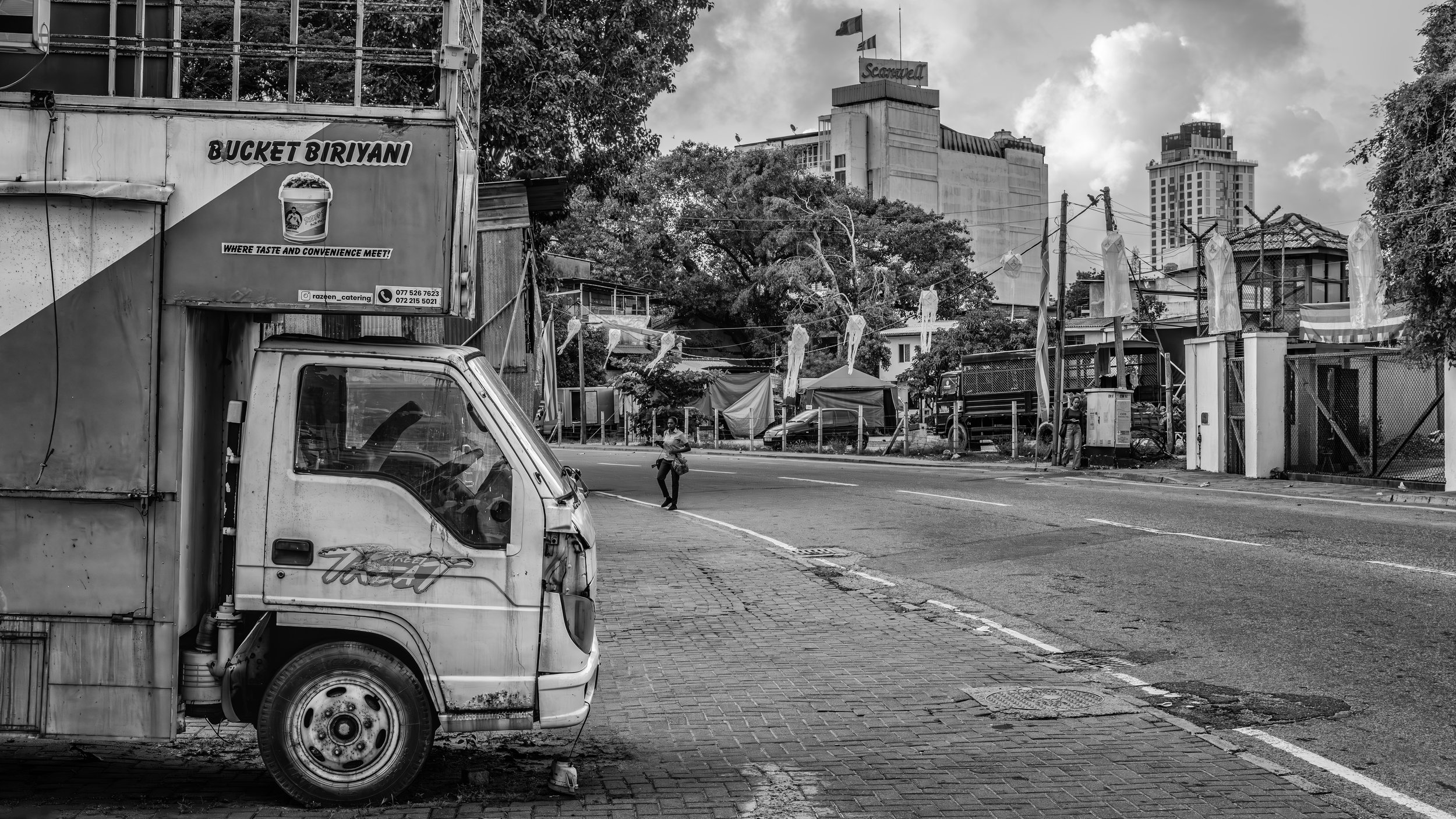 A black and white street scene featuring parked trucks and a pedestrian crossing the street. There are buildings, trees, and flags in the background, with some tents or structures along the sidewalk.