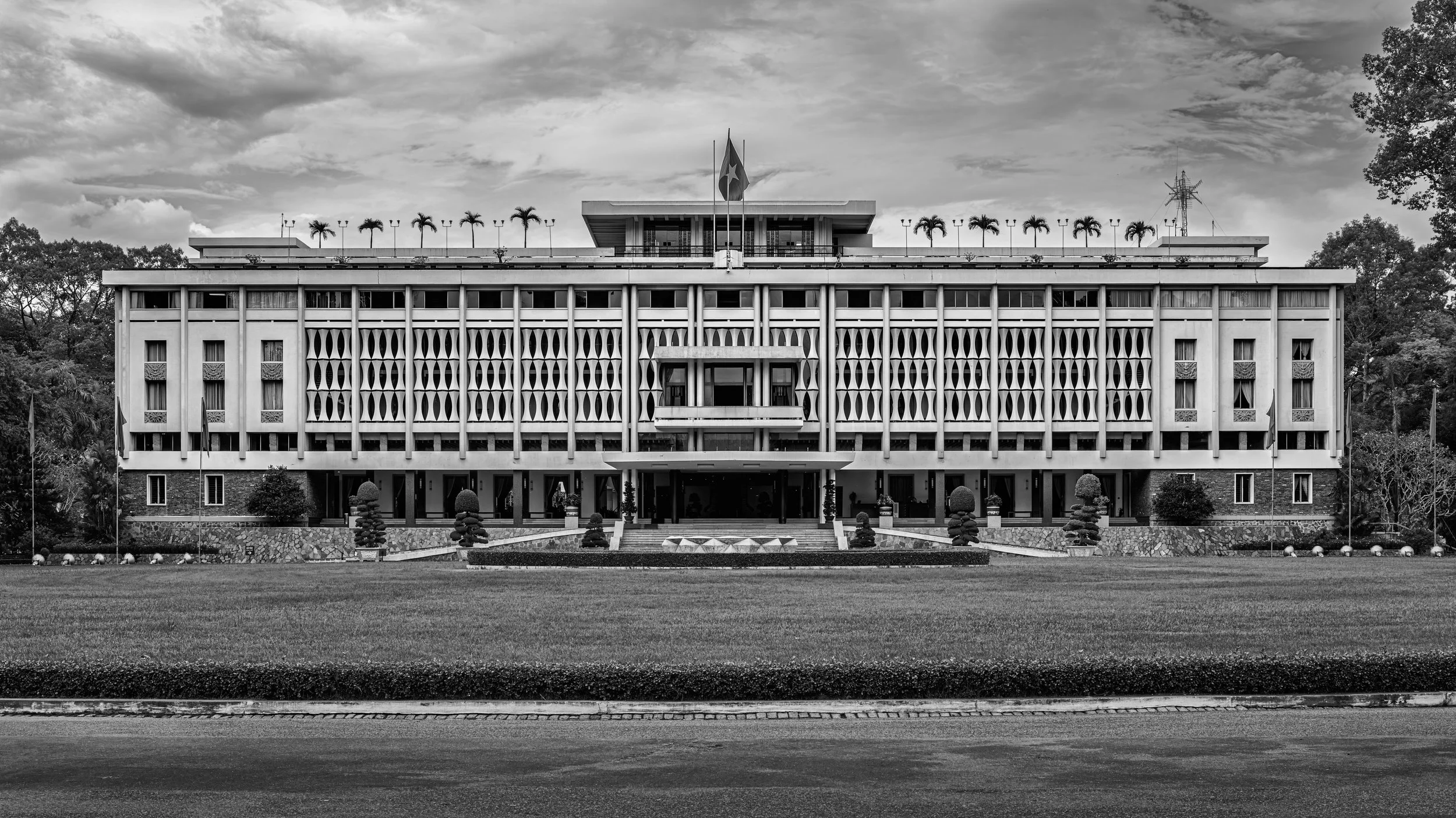 A large government building with a flagpole and two flags on top, palm trees along the roofline, and a well-maintained lawn in front, black and white photo.