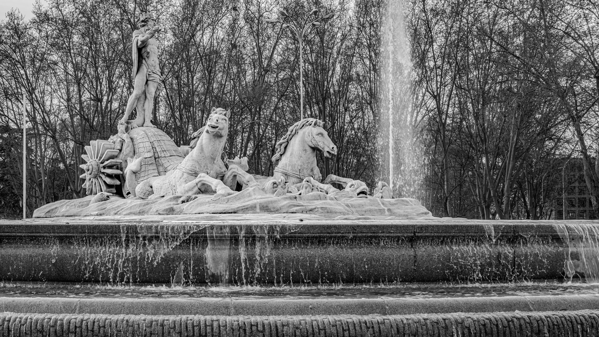 A black and white photo of a large ornate fountain with multiple statues depicting mythological figures and animals, with water flowing into a lower basin.