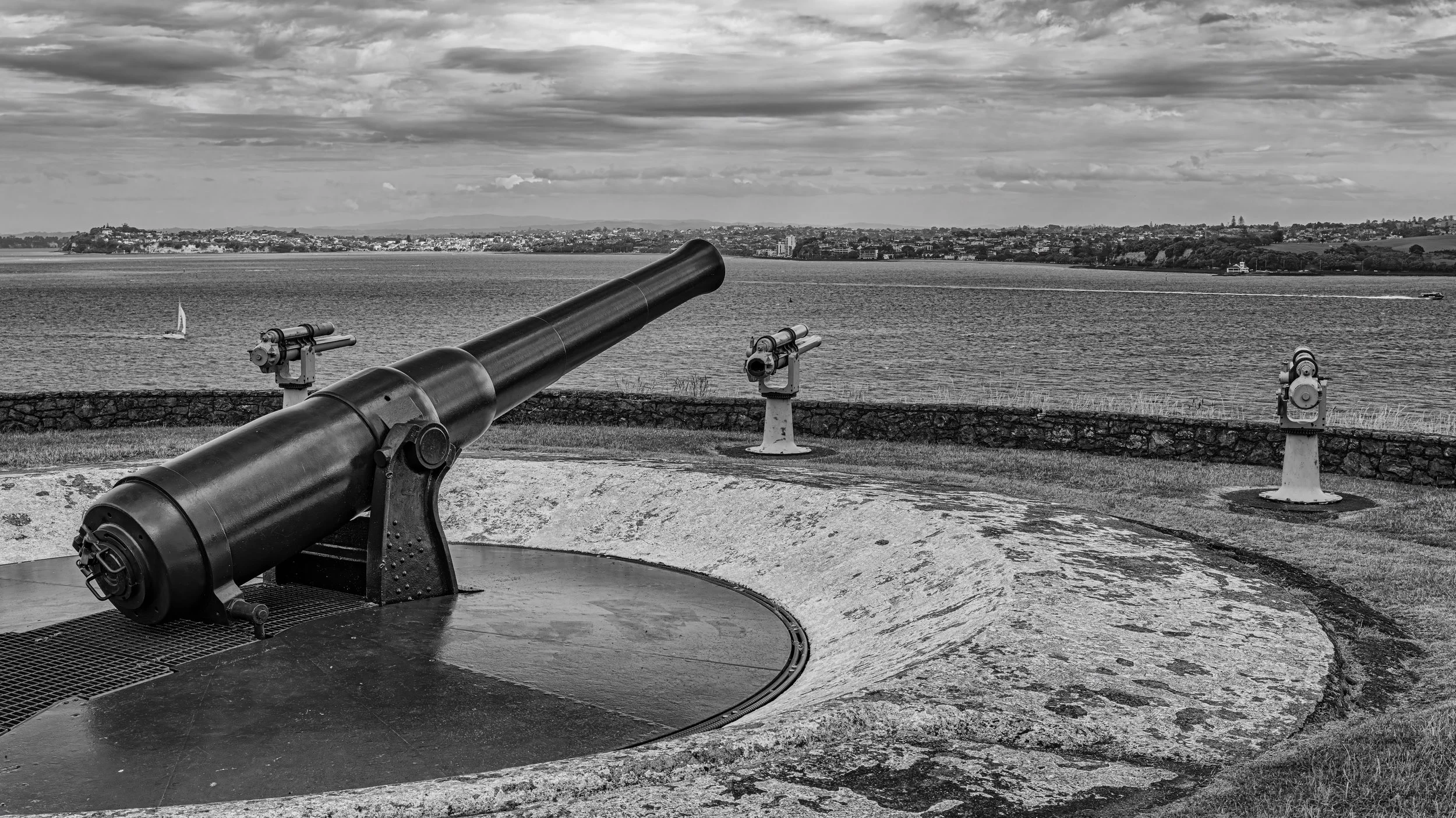 A large cannon overlooking a body of water with islands or land in the distance, under a cloudy sky.