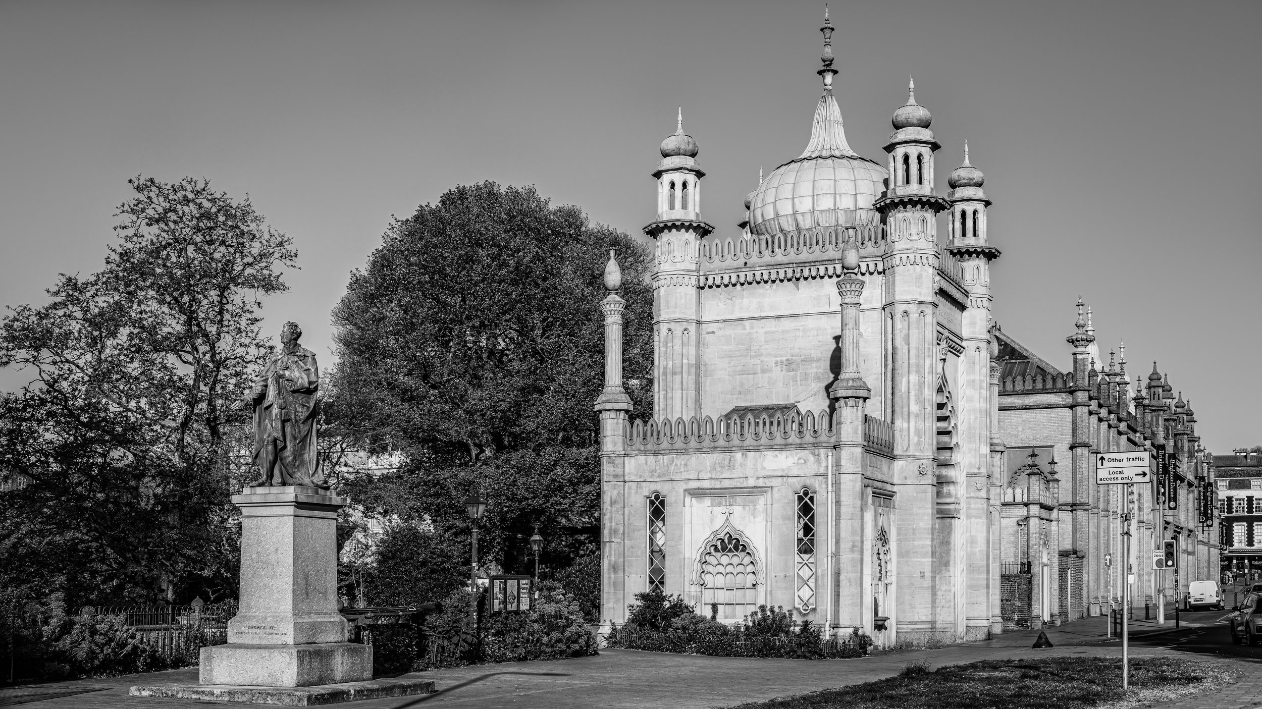 A historic city scene with a statue of a man in a robe on a pedestal, surrounded by trees. A Gothic-style church with domes and spires is visible in the background, with cars and street signs nearby.