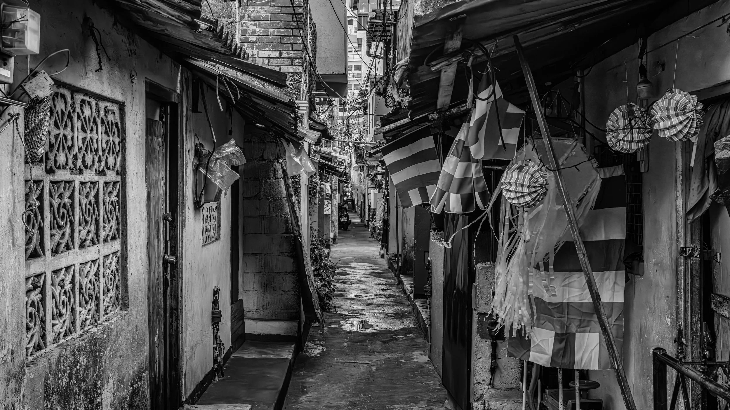 Narrow alleyway between buildings with hanging flags and decorations, some string lights, and a wet pavement.