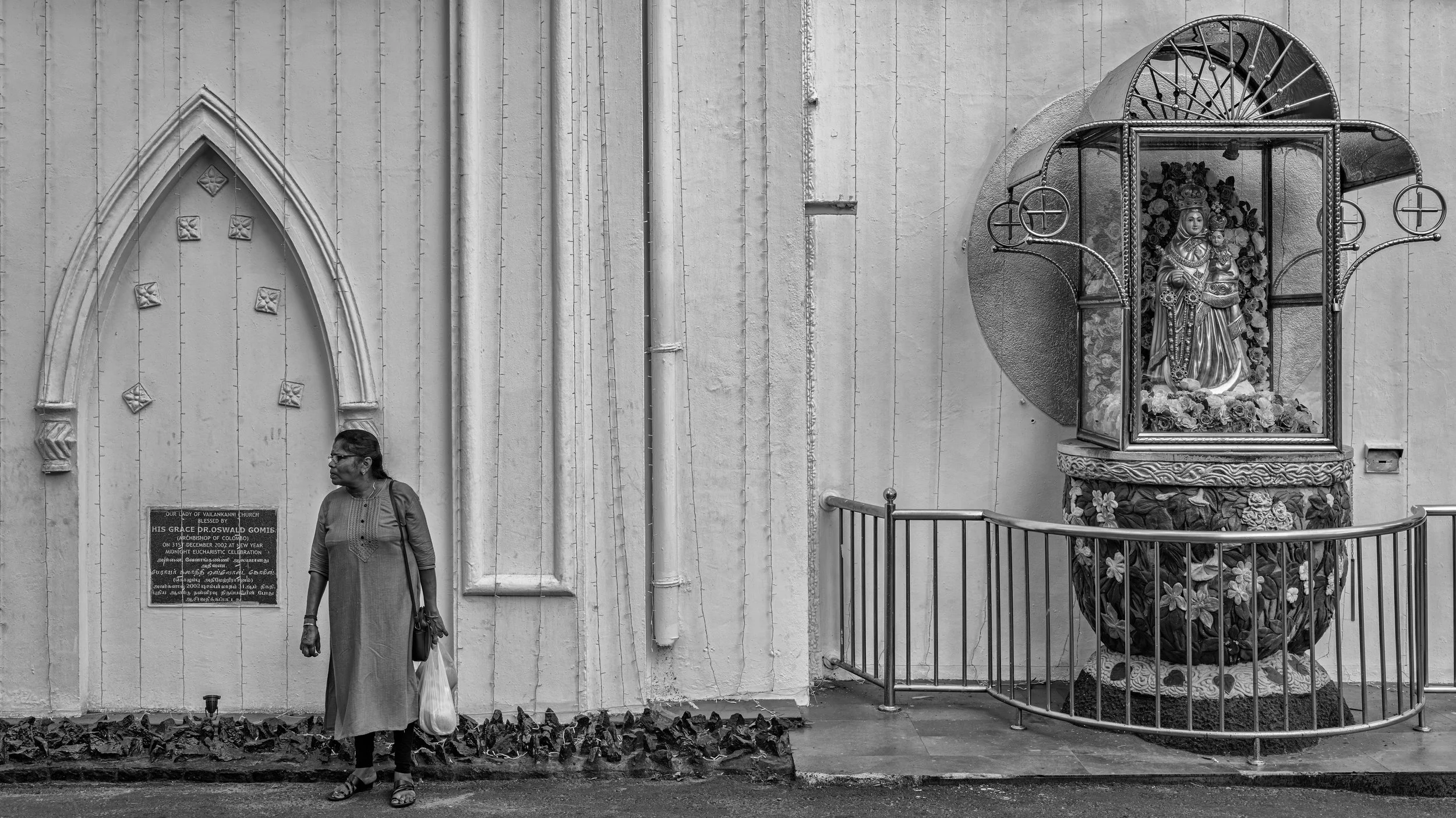 A woman in traditional attire standing outside a church, next to a religious shrine with a statue of the Virgin Mary holding baby Jesus, surrounded by flowers, enclosed in a decorative metal casing.