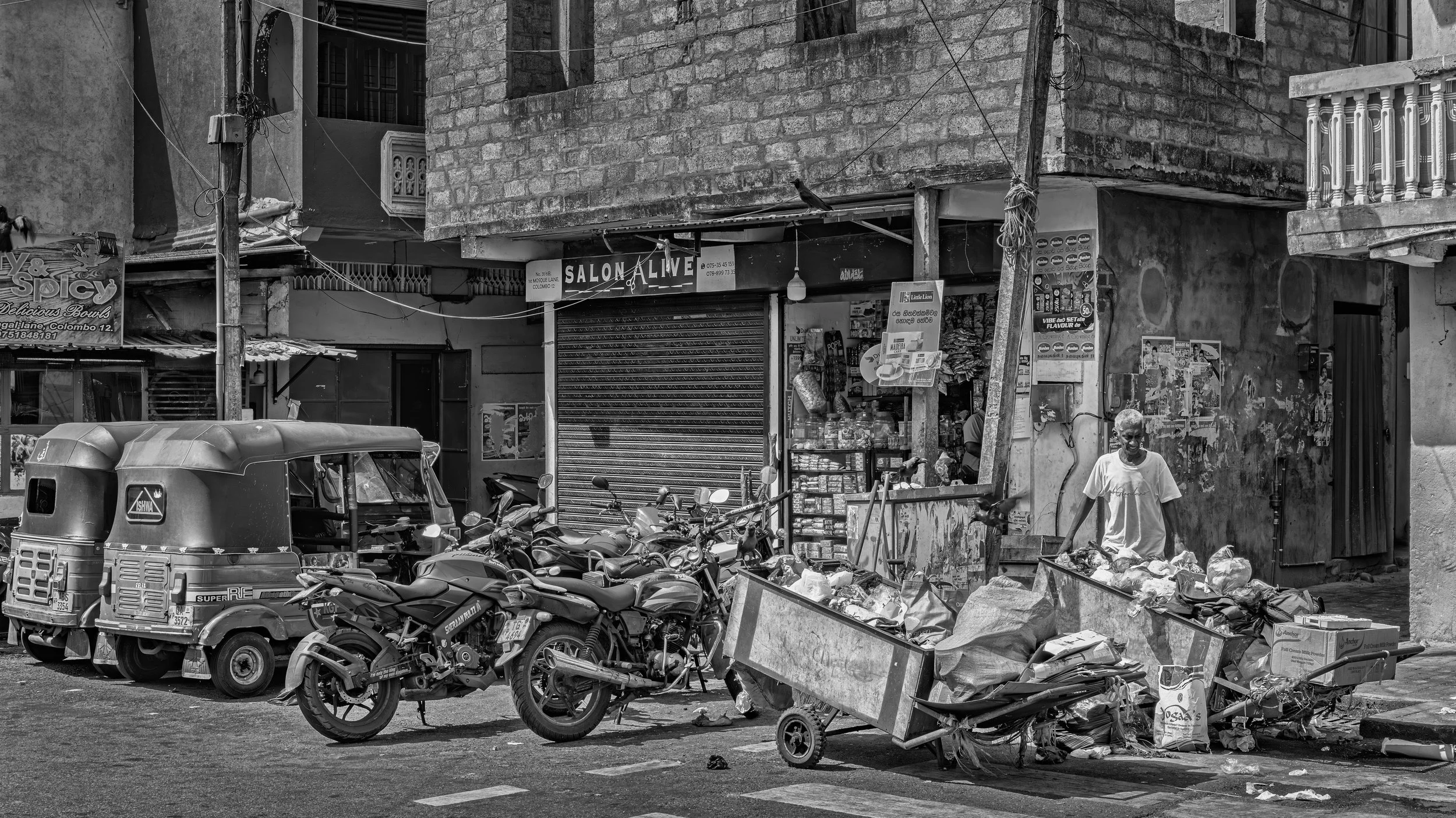 Street scene with parked motorcycles and an auto rickshaw in front of small shops, one sign reads 'Salon Alive'. A man with gray hair and wearing a white t-shirt stands near a cart filled with bags, in a neighborhood with brick buildings and wires.