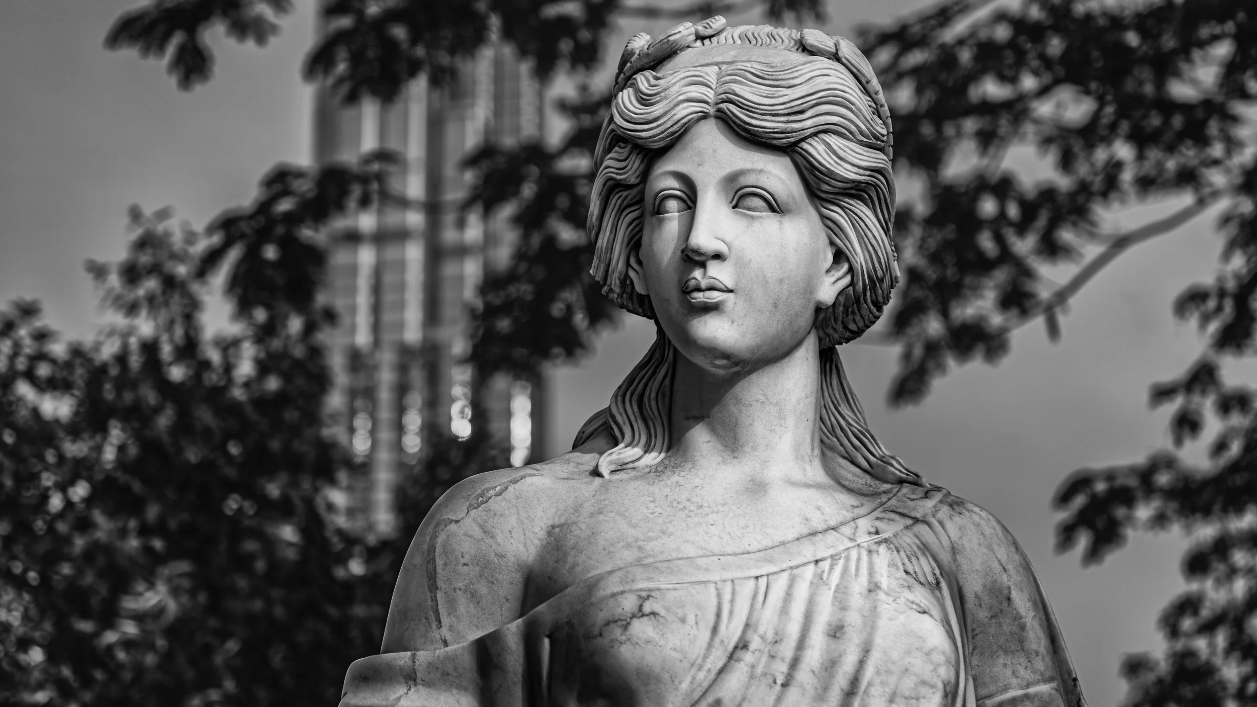 Black and white photo of a marble statue of a woman with long hair, with trees and a building in the background.