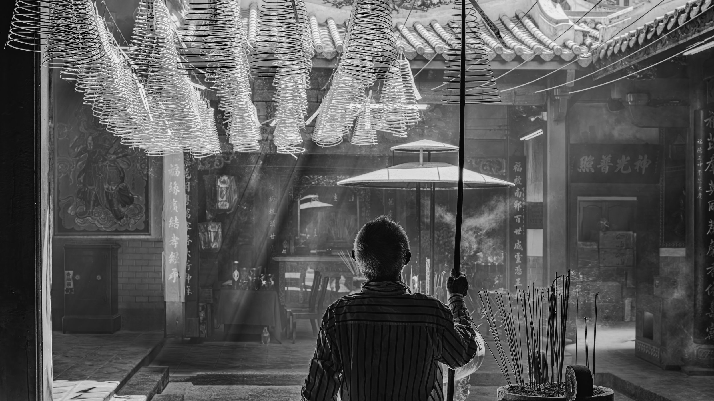 A boy holding an umbrella and a stick, standing in front of a temple or traditional building with hanging lanterns and Asian script on signs and banners, backlit by sunlight. Saigon, Vietnam.