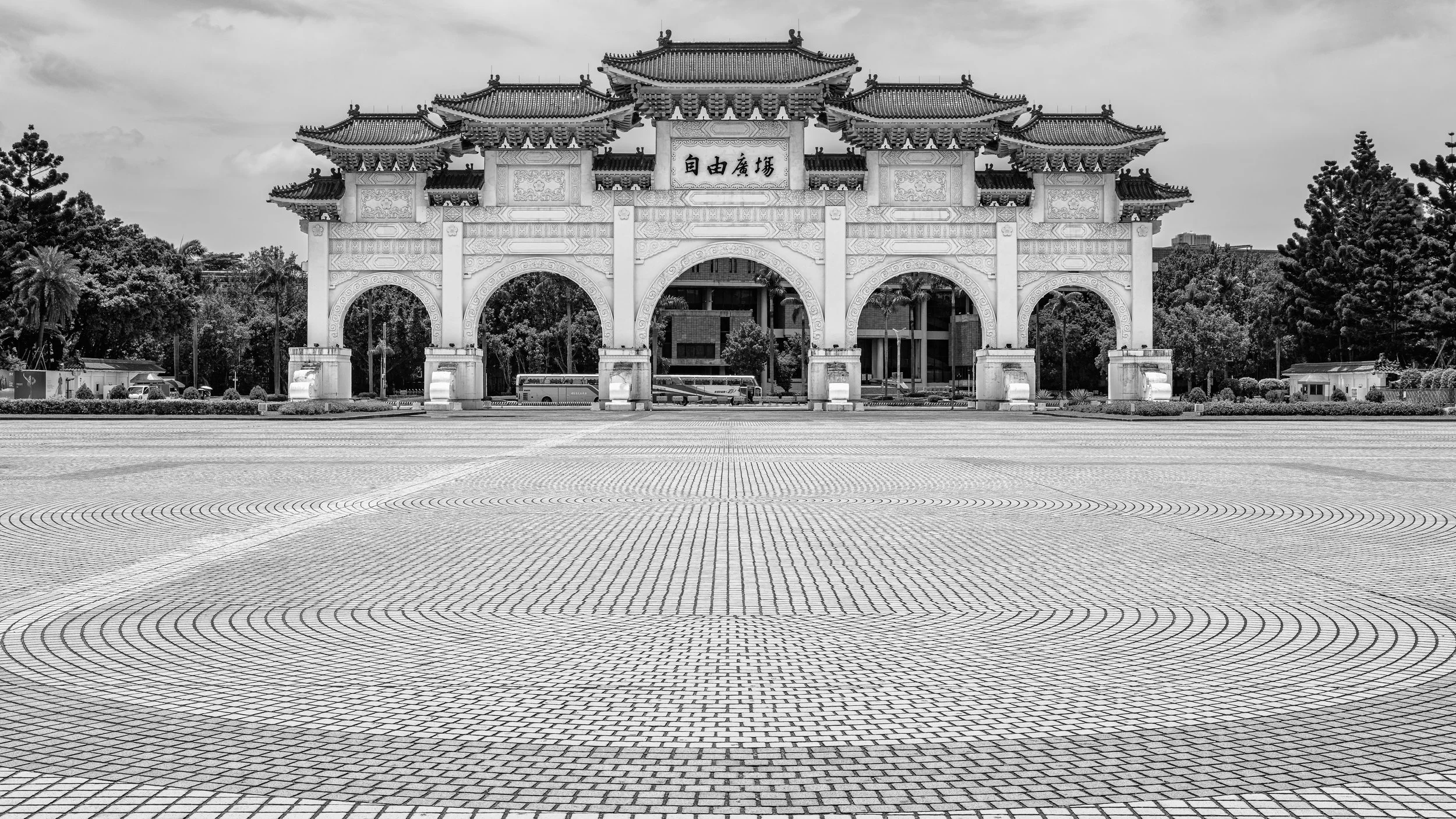 Traditional Chinese-style gateway with three large archways, decorative roof, and intricate designs, set against trees and buildings with a tiled ground in the foreground.