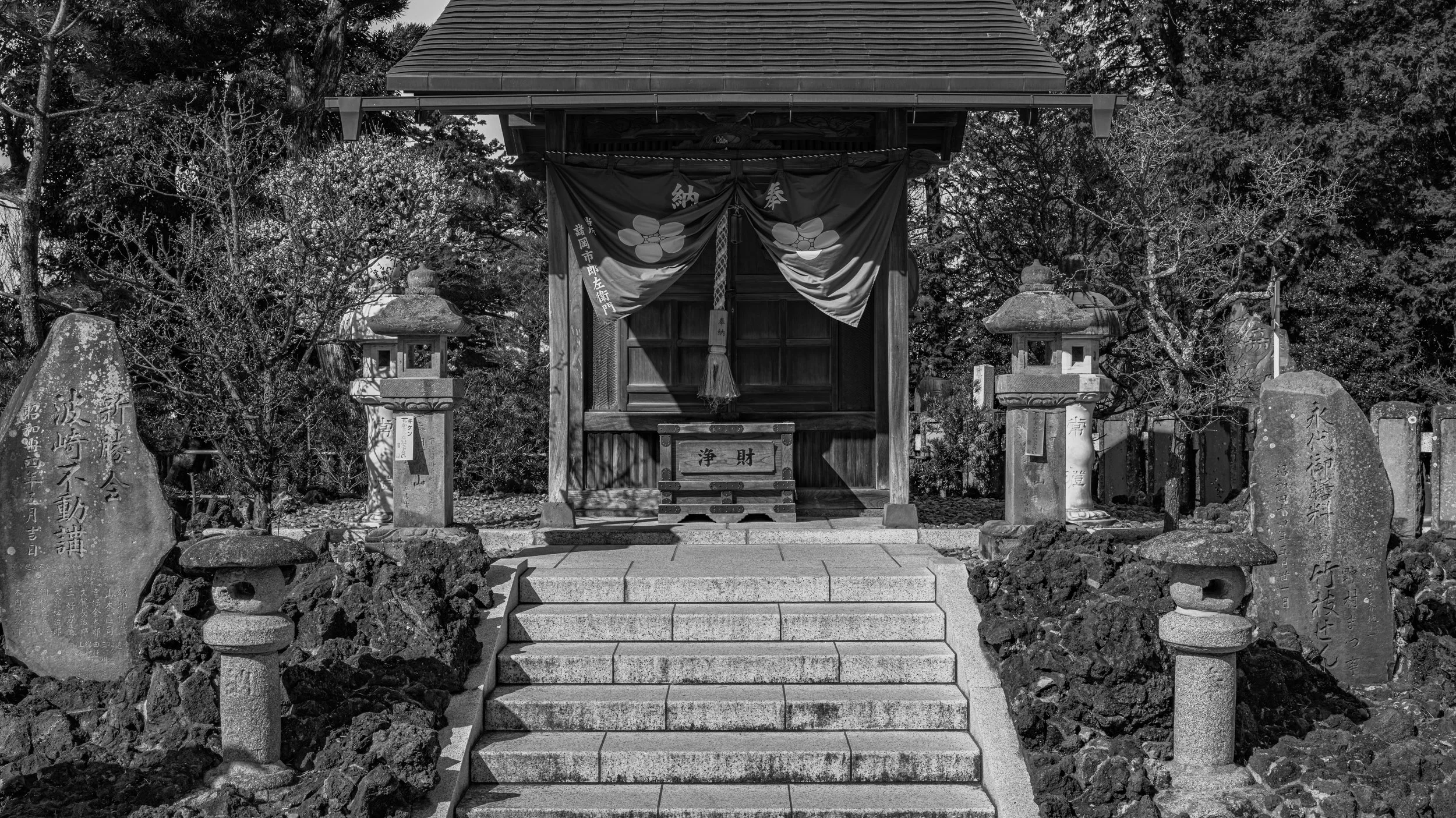 A small traditional Japanese shrine with stone steps leading up to it, surrounded by stone lanterns and inscriptions on stone tablets, with trees in the background.