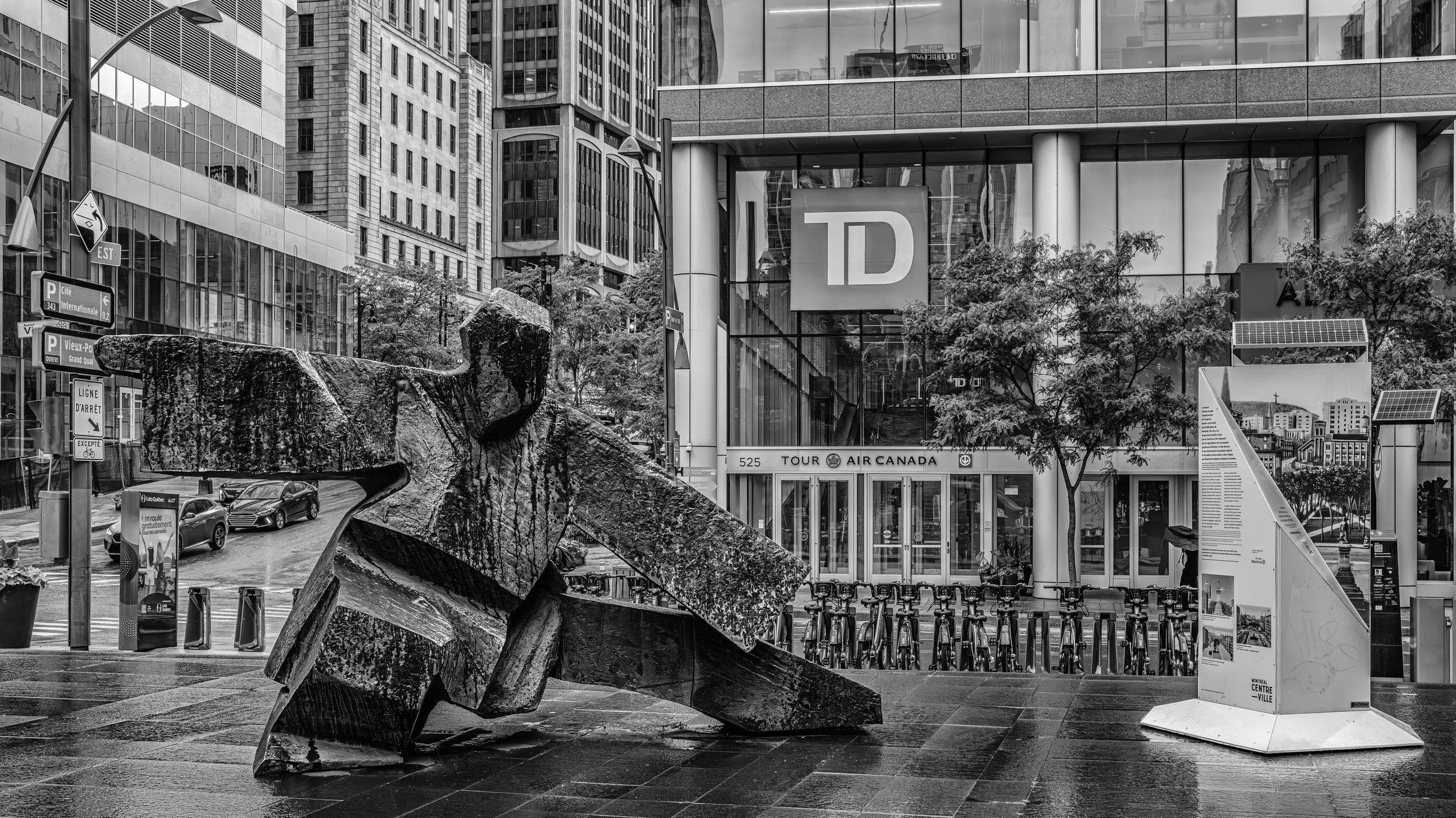 Black and white cityscape with modern glass buildings, trees, and a large abstract rock sculpture in the foreground.
