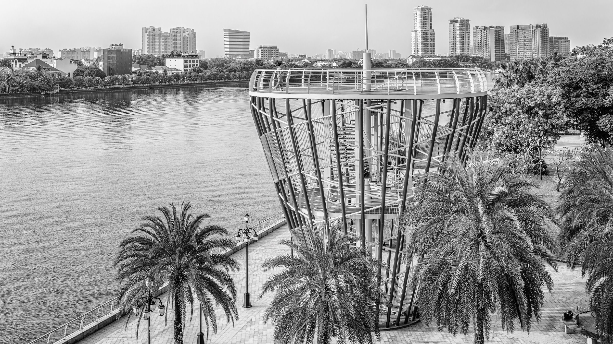 Black and white photo of a modern spiral observation tower next to a body of water with a city skyline in the background, surrounded by palm trees.