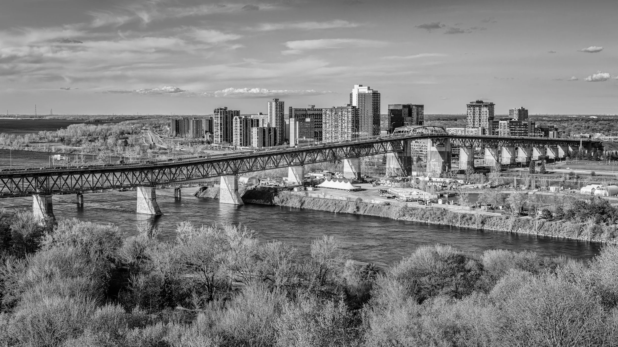 Black and white cityscape with a river, bridge, and high-rise buildings under a partly cloudy sky.