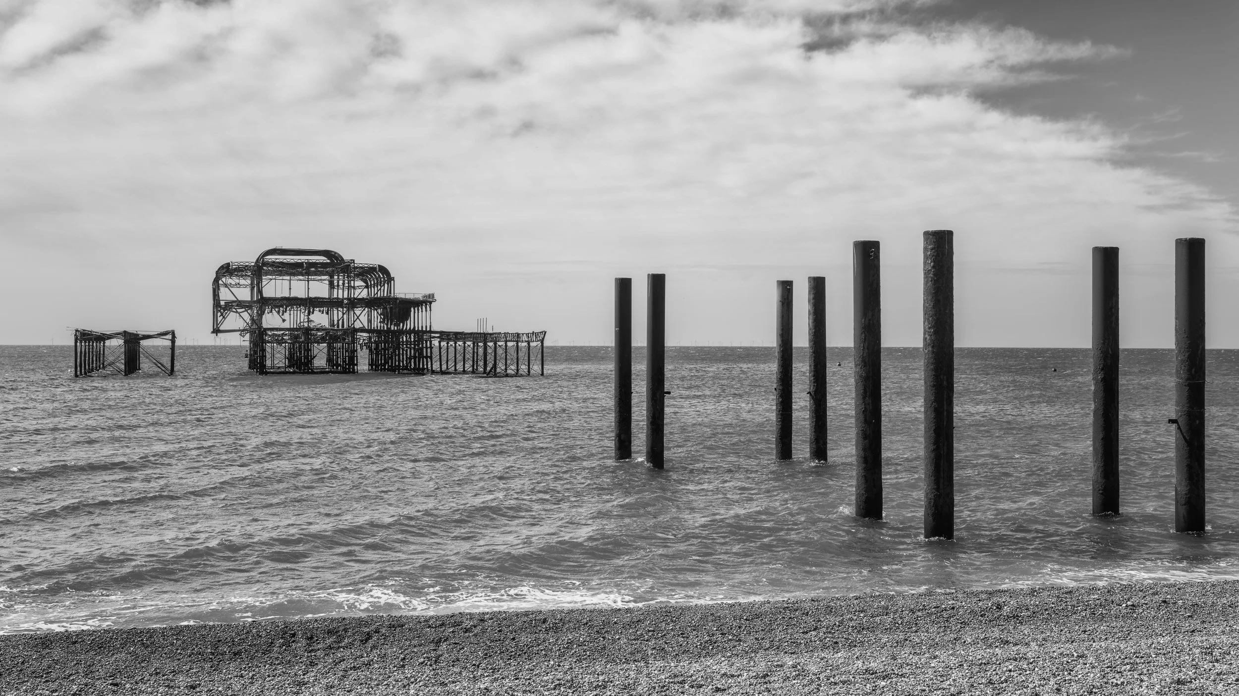 Black and white photo of a partially sunken, rusted amusement park ride in the ocean, with a few upright pillars in the water and a cloudy sky overhead.