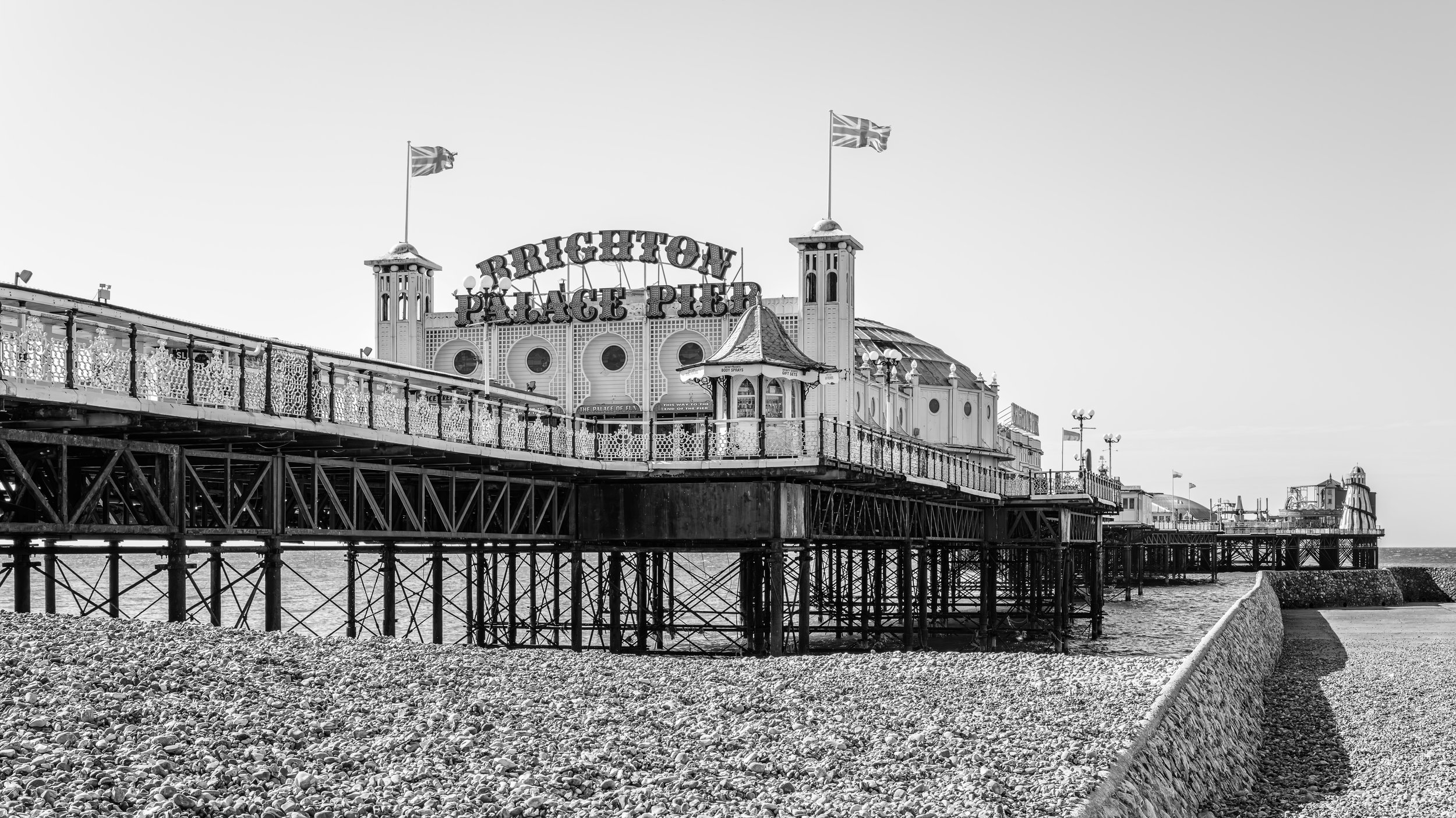 Black and white photo of Brighton Palace Pier with flags on top, a walkway with decorative railings, and a rocky beach in the foreground.
