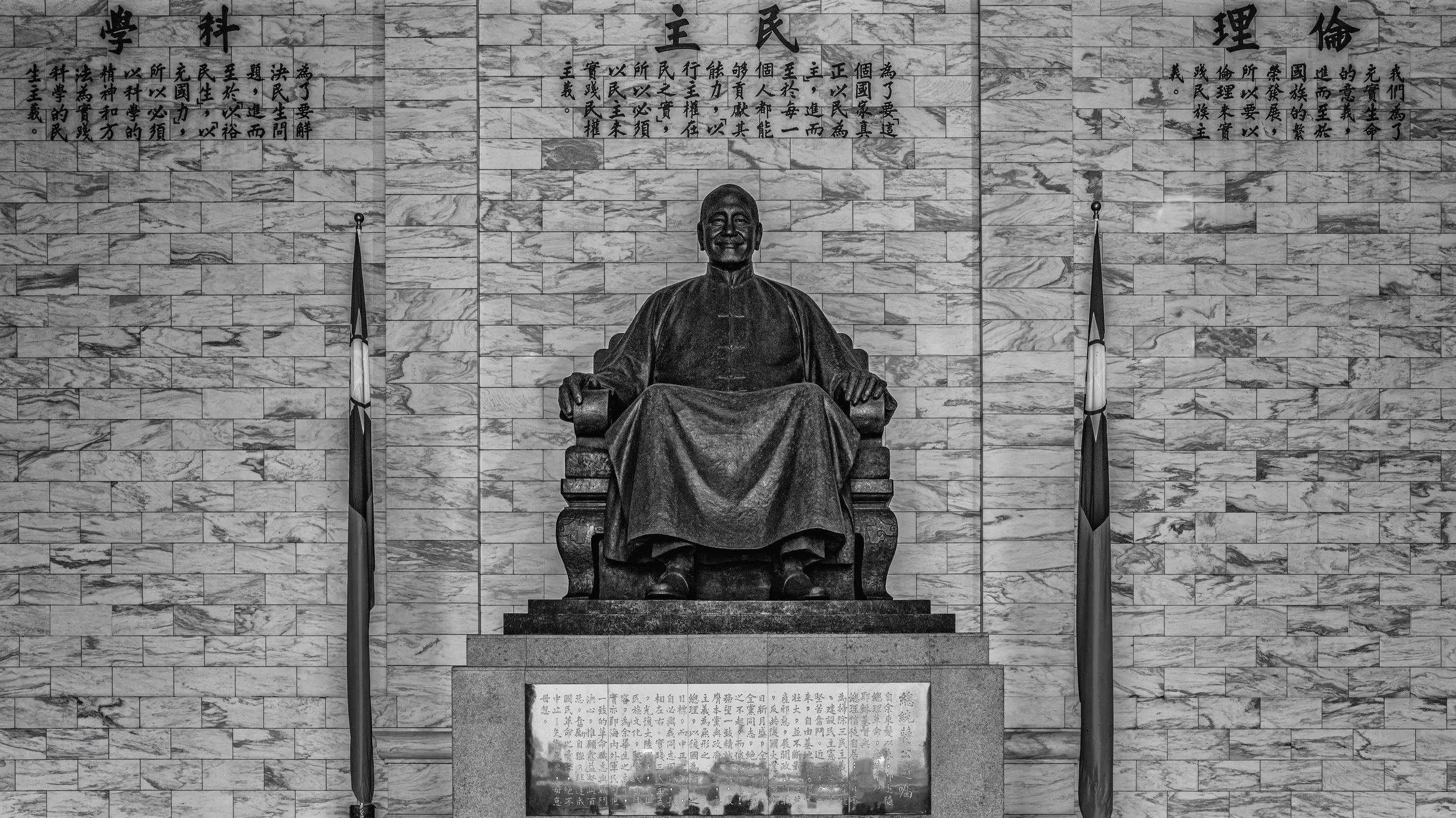 A bronze statue of Sun Yat-sen seated on a throne, located in front of a marble brick wall with Chinese characters above. Two flags are positioned on either side of the statue, and there is an inscription plaque with Chinese text below.