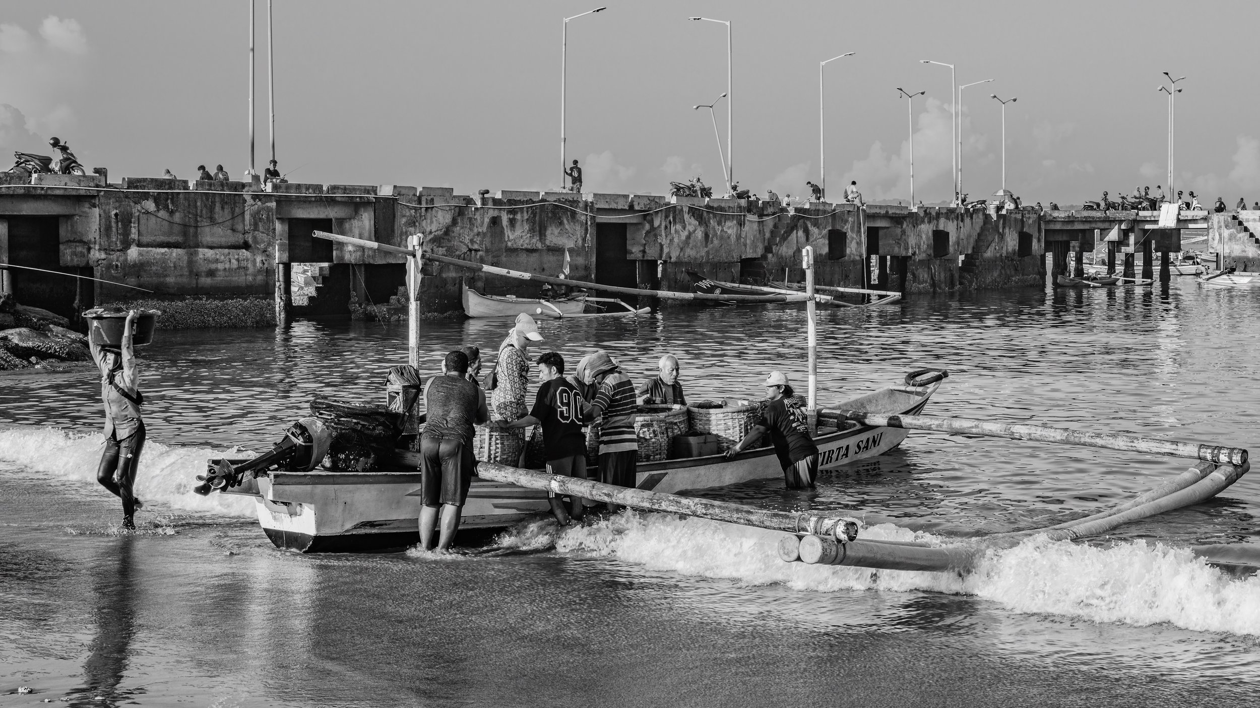 People on a boat and on the shore near a concrete pier with streetlights, some fishing and some walking or sitting, in a black and white photo.