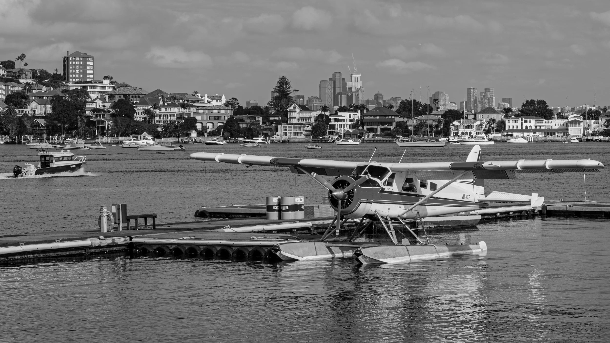 A harbor with boats and a seaplane floating on water, with a city skyline featuring tall buildings and houses on a hillside in the background, under a partly cloudy sky.