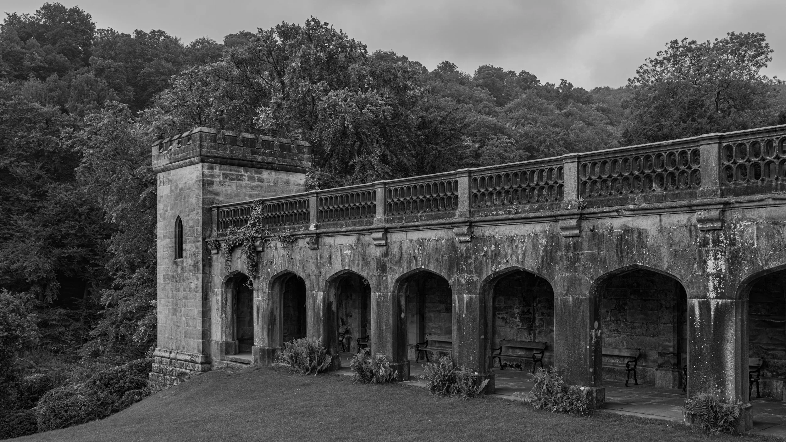 Black and white photo of an old stone arches bridge with a decorative stone wall on top, surrounded by trees and a garden in the foreground.