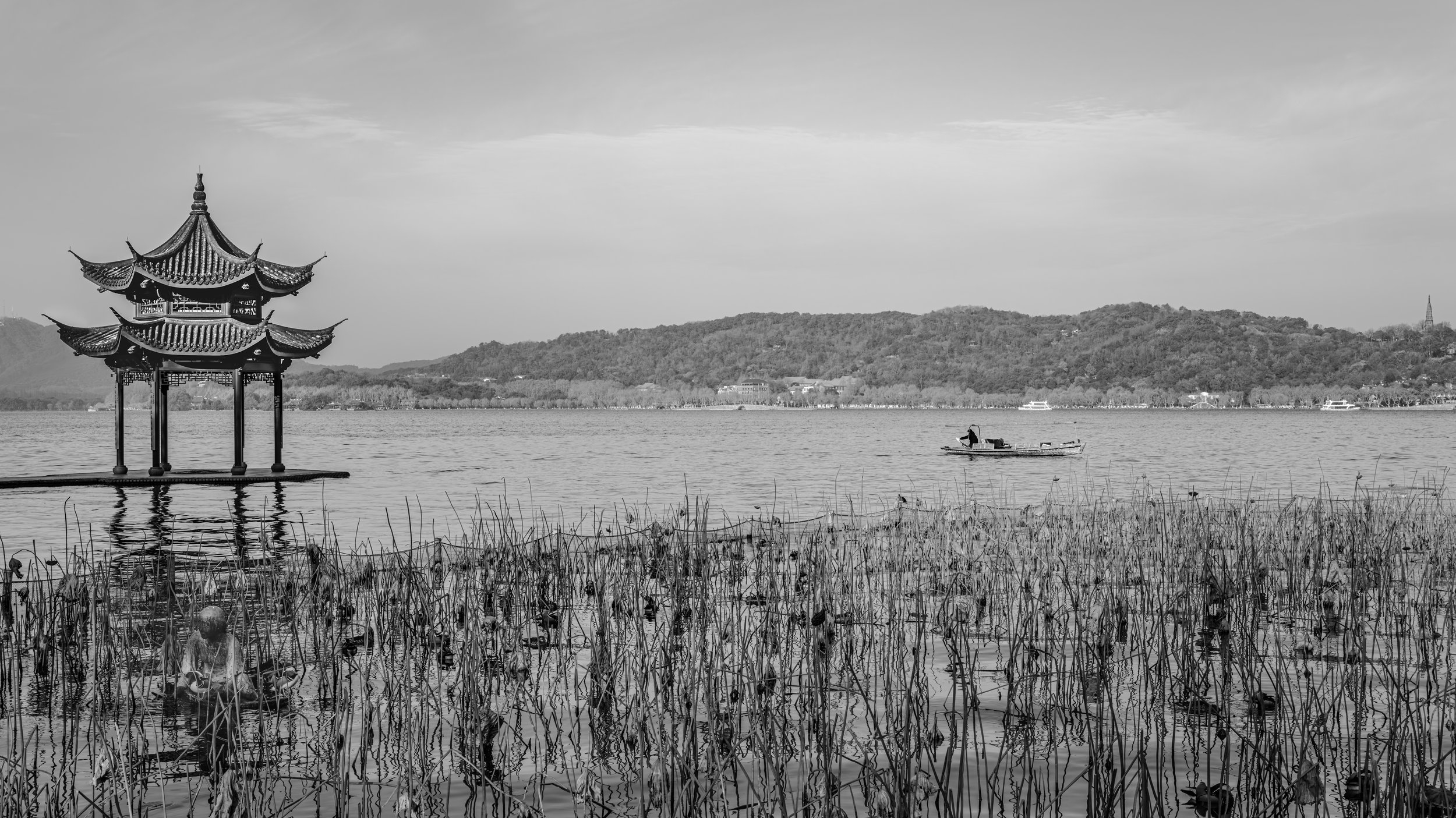 A traditional Chinese pagoda on stilts over a lake, with a small boat and distant hills in the background, black and white photograph.