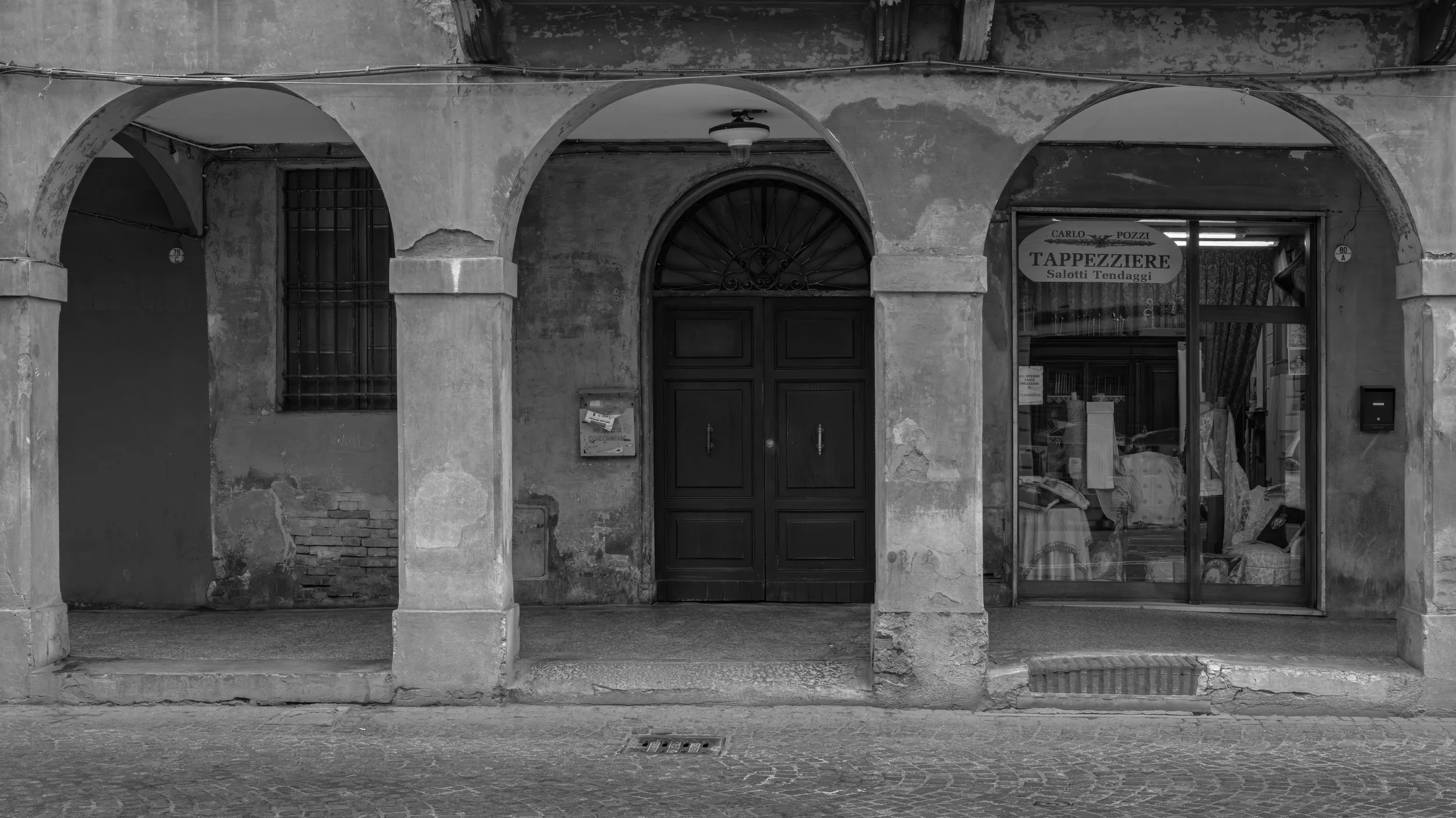 An old building facade with three arched openings, one of which is a closed door, another an empty archway, and the third a shop with a sign that reads 'Tappezziere Salotti Tendaggi'. The building shows signs of wear with peeling paint and cracks. Th