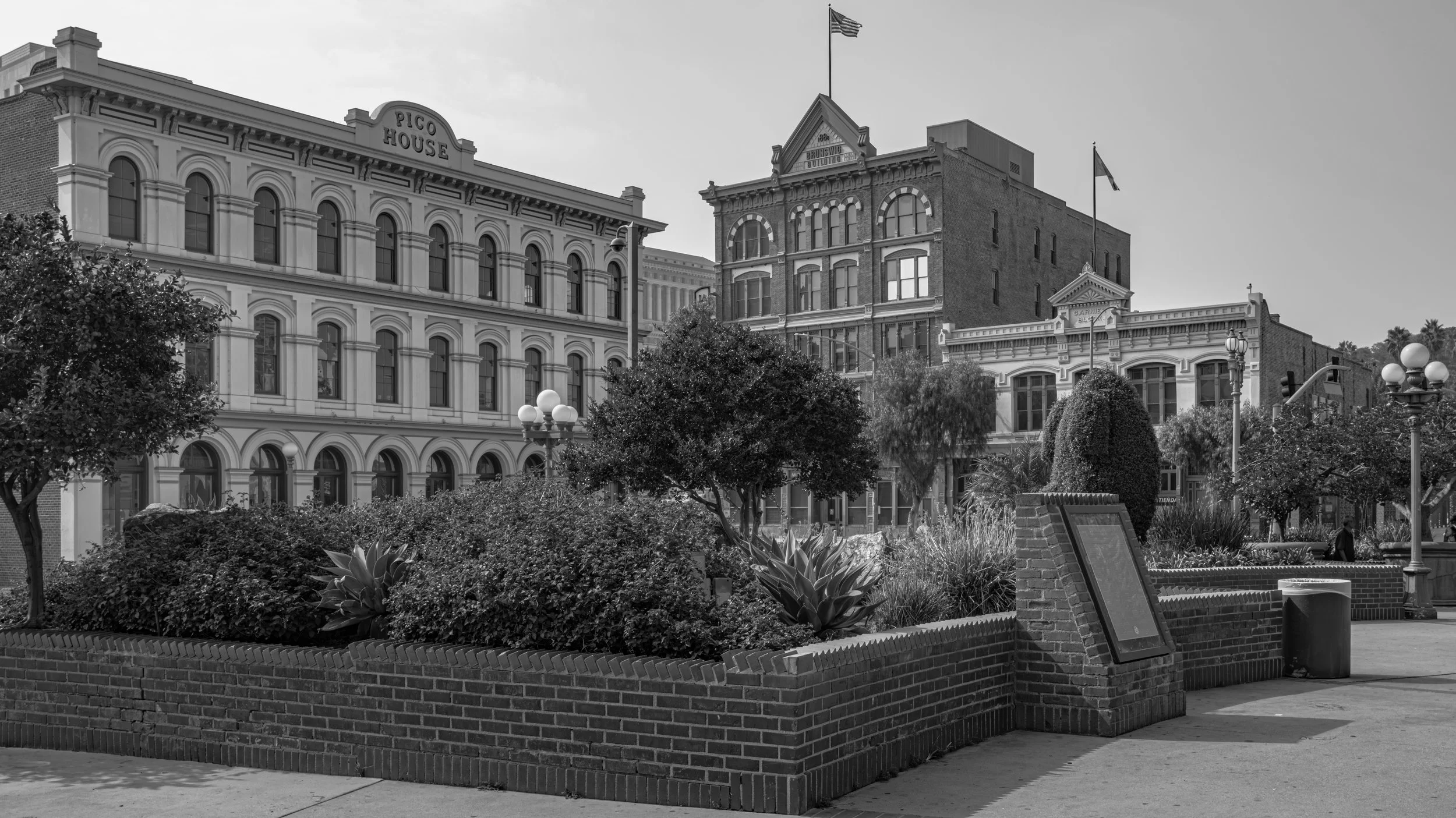 A city street scene in black and white with historic buildings, trees, and street lamps, including a sign for Pico House and flags flying on top of the buildings.