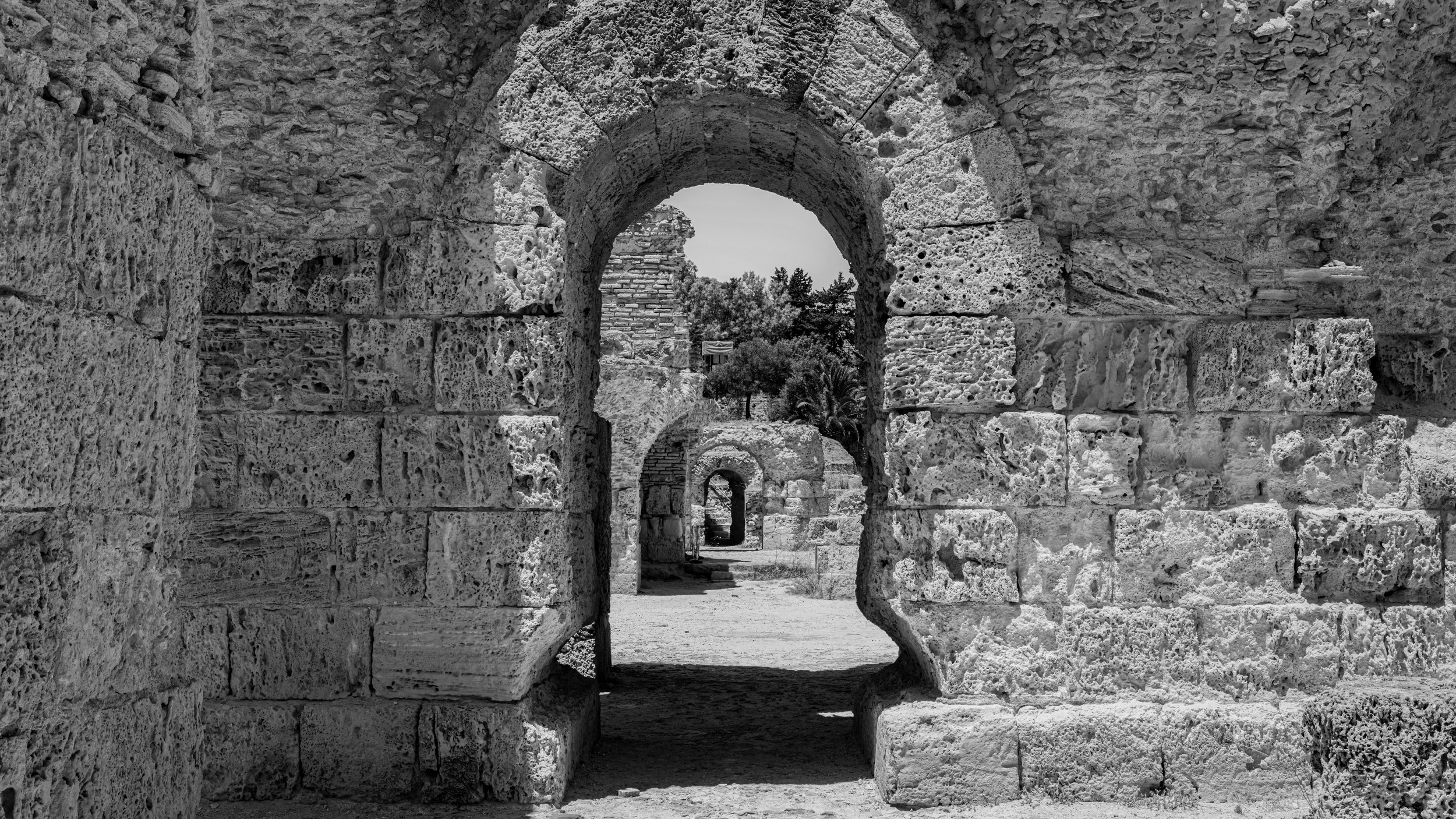 Black and white photo of ancient stone ruins with several arches and a pathway, trees in the background.