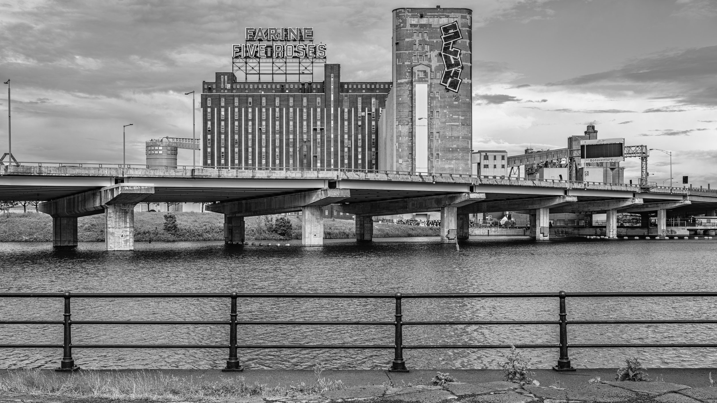 Black and white photo of a cityscape with a river in the foreground, an overpass bridge, and a large industrial building with signage that reads "FARINE FIVE ROSES." The building has a cylindrical silo with graffiti on it, and the sky is cloudy.