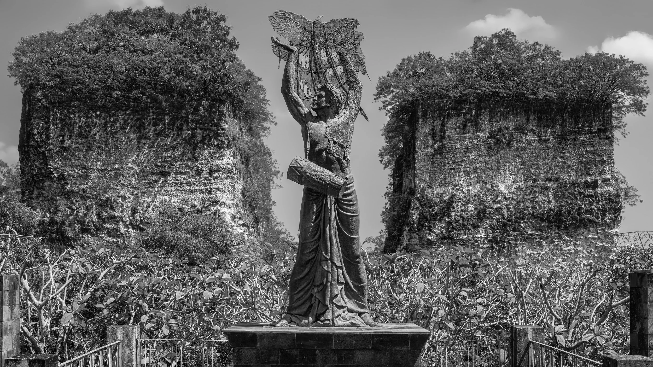 Black and white photo of a statue of a woman holding a butterfly-shaped sculpture above her head, surrounded by trees and bushes.