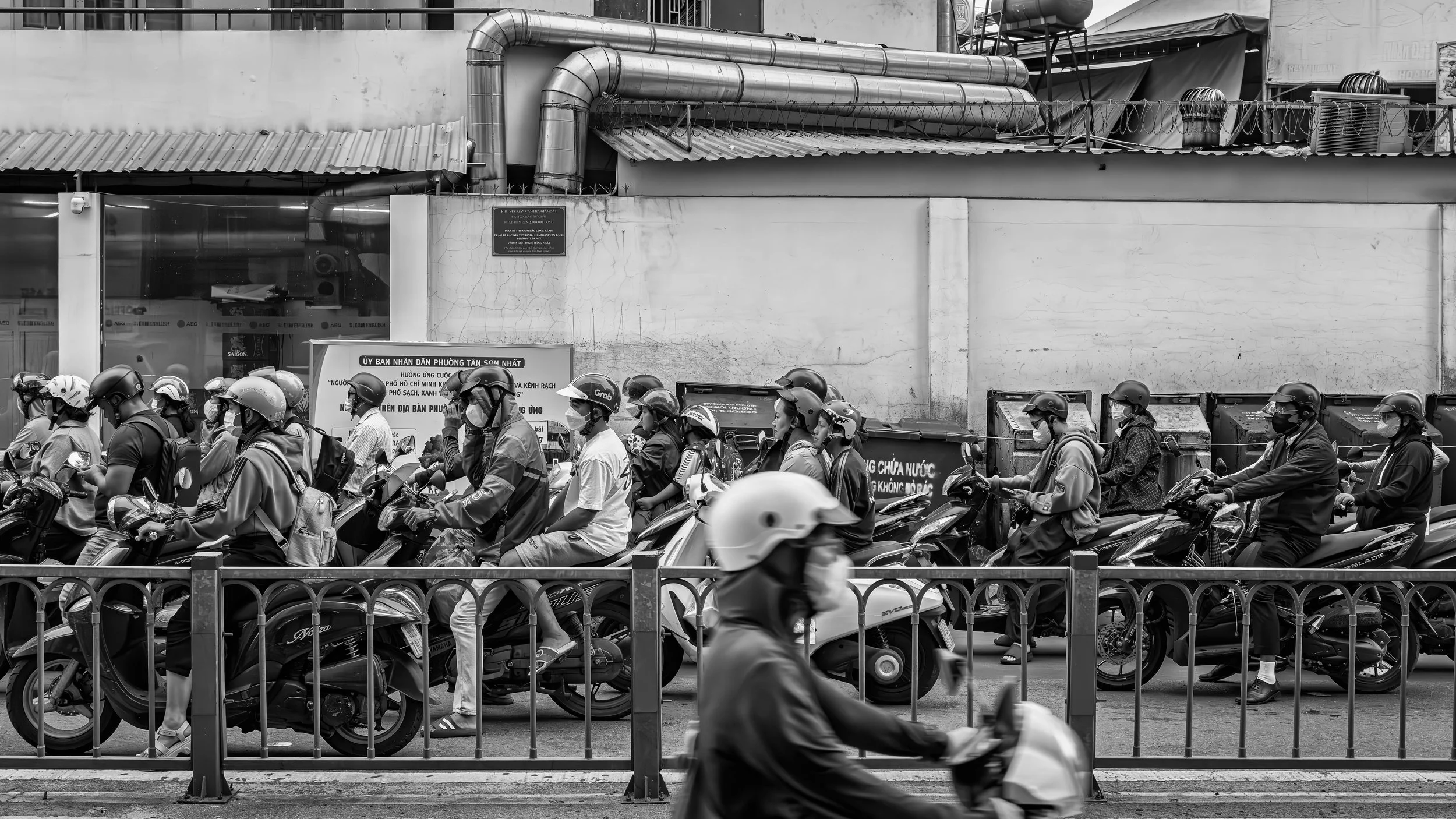 A large group of motorbikes and scooters lined up on the street with riders wearing helmets, some with masks, moving in the same direction. The background shows a wall with some pipes and a sign in Vietnamese.