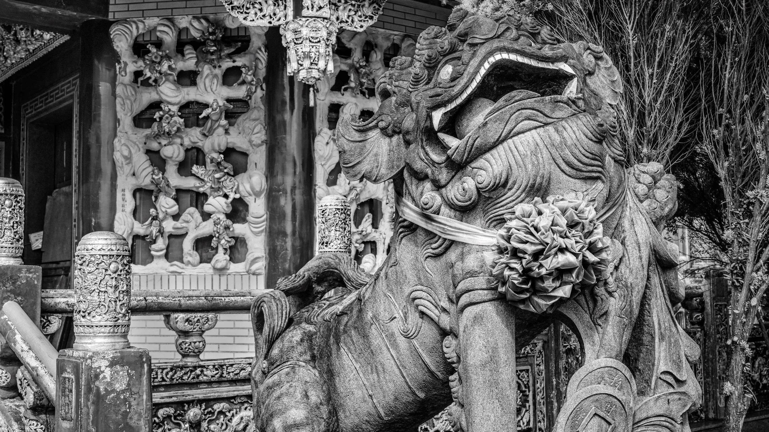 A detailed stone statue of a mythical lion, possibly a Chinese guardian lion, with intricate carvings and a ribbon or cloth tied around its neck, situated outdoors. In the background, there are ornate wood carvings showcasing figures and traditional 