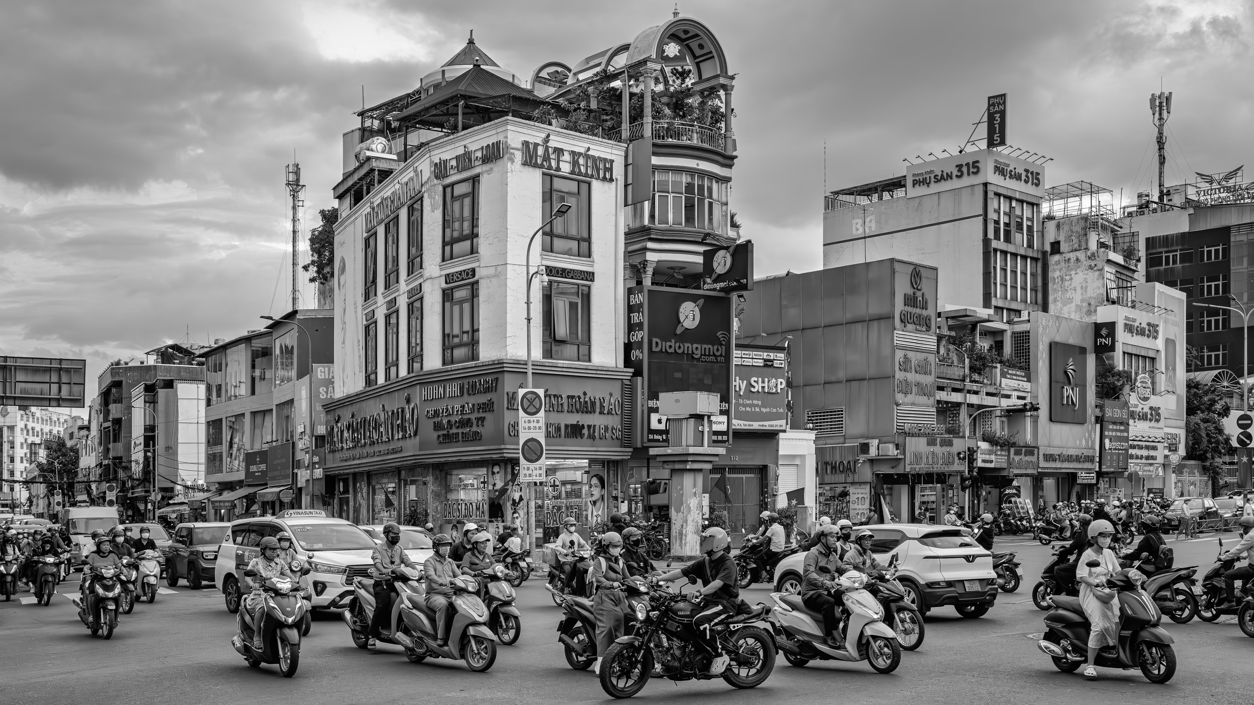 A busy city street with many motorbikes and cars at a crosswalk. Tall buildings line the street with various signs and advertisements, some in Vietnamese. The sky is cloudy.