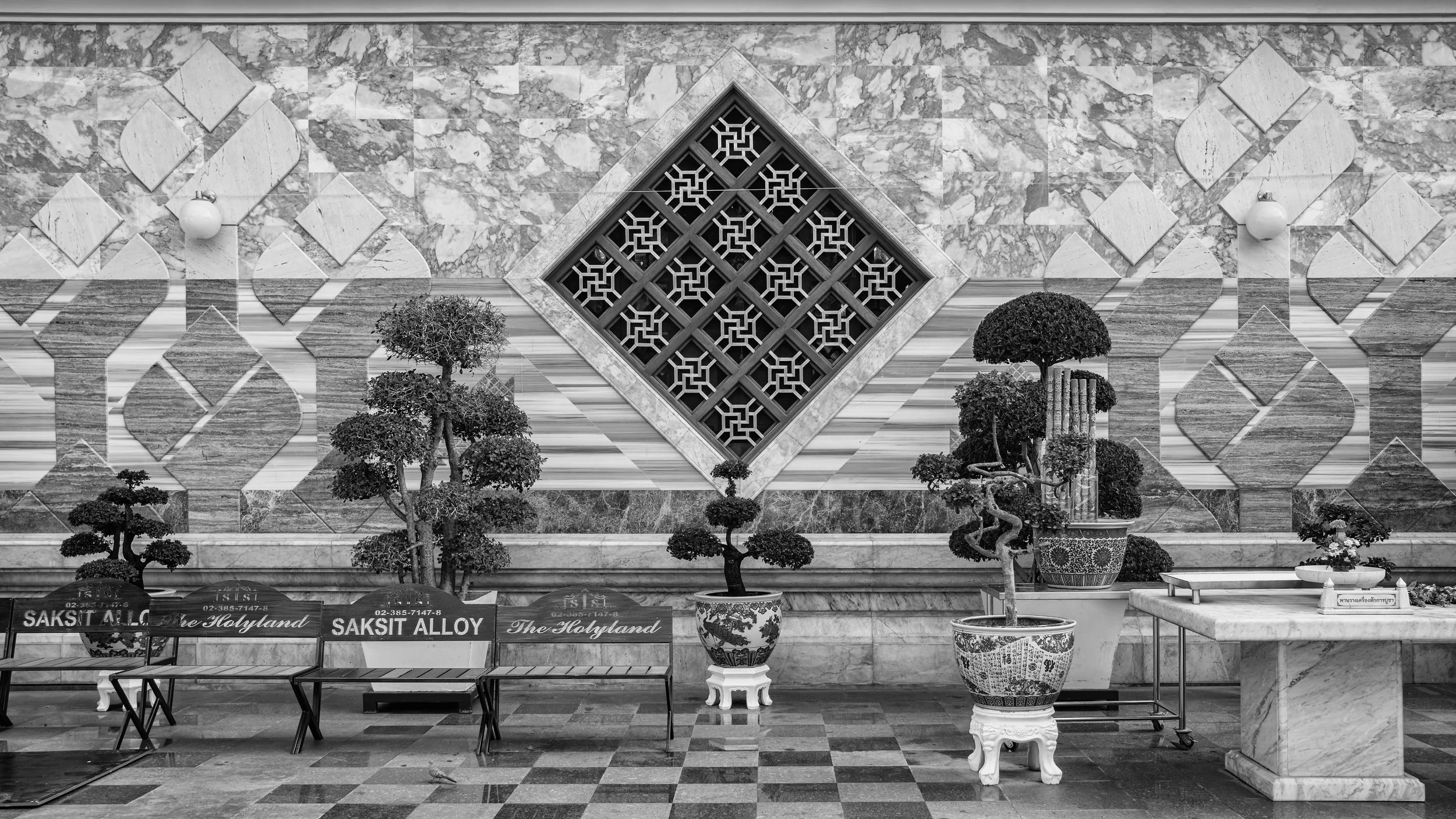 Black and white photo of a decorative interior wall with a diamond-shaped lattice window, surrounded by marble tiles. There are small ornamental trees and potted plants in front of the wall, with a bench and flowers below. Bangkok. Thailand.
