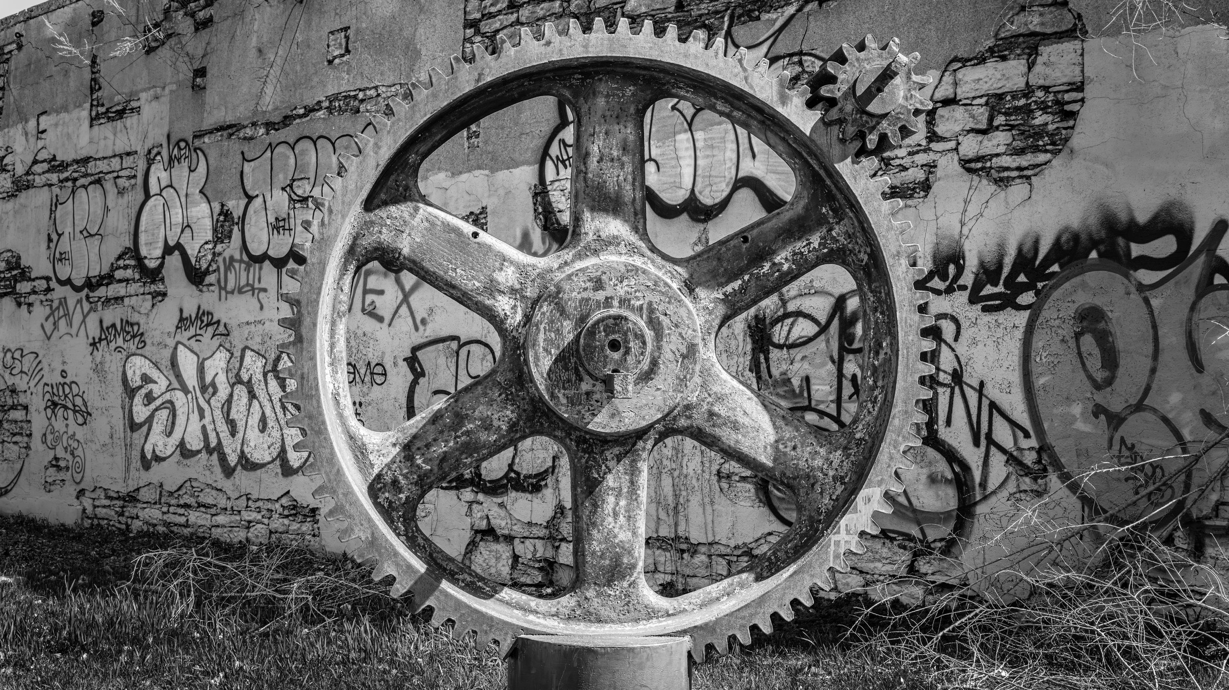 A large, weathered gear mounted on a stand in front of a graffiti-covered brick wall with some plants and grass at the base.