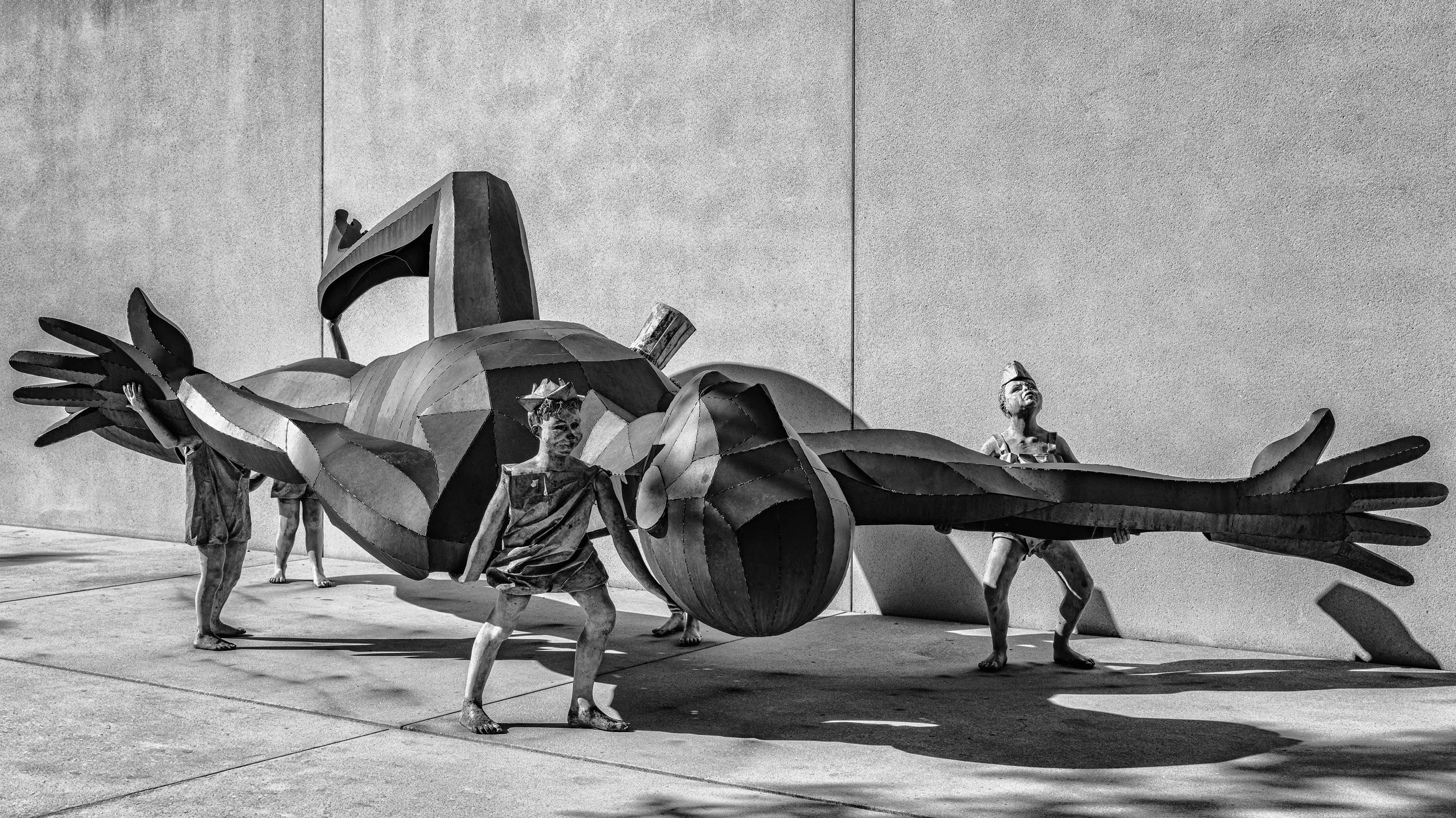 Black and white sculpture of large hands, body, wings, and head of a fly, with children standing around and touching it, against a plain wall.