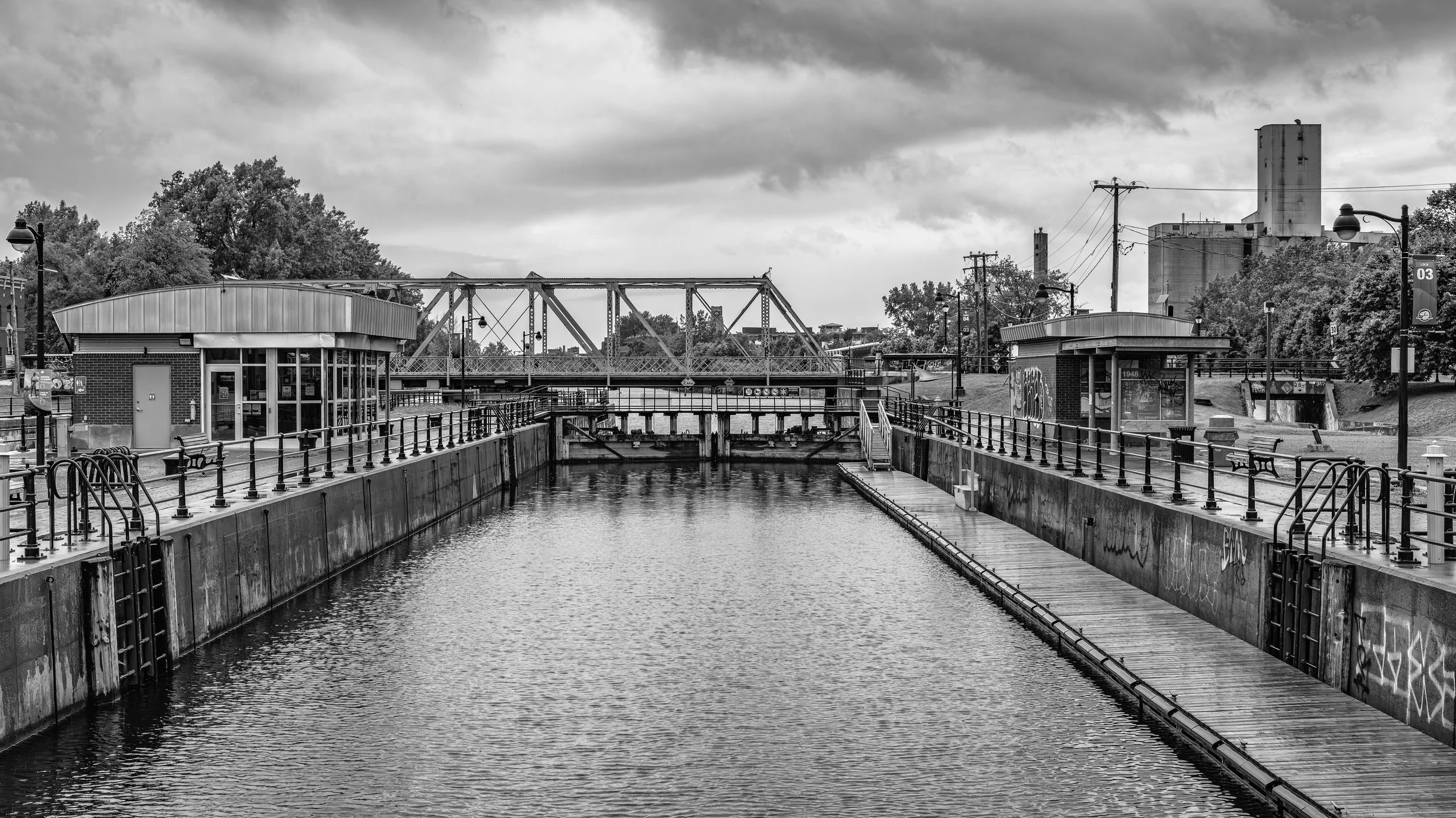 A black and white photo of a canal with lock gates and walkways, surrounded by trees, buildings, and lampposts, under a cloudy sky.