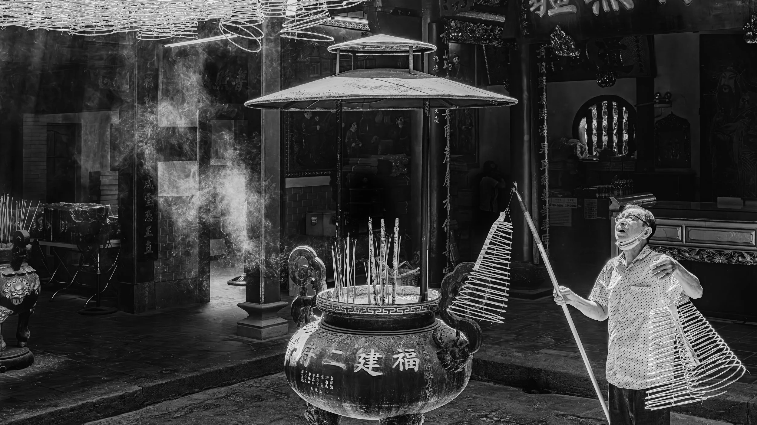 A man wearing glasses and a face mask is holding a fan-shaped incense holder with smoke rising from it inside a traditional Chinese temple with various statues, banners, and incense sticks.