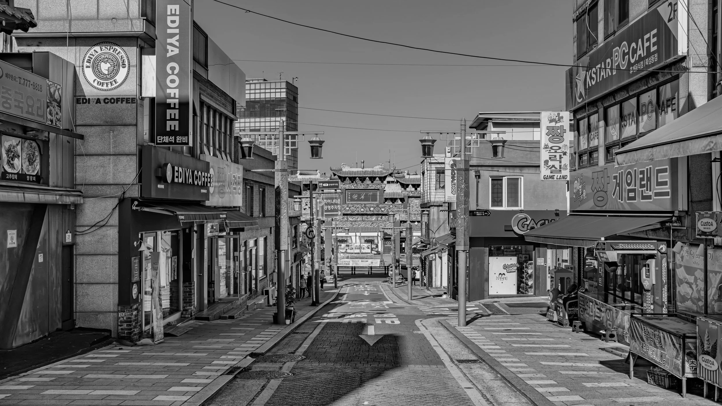 A black and white photograph of a city street with various storefronts, signs, and a traditional ornate gate in the background, indicating an Asian influence.