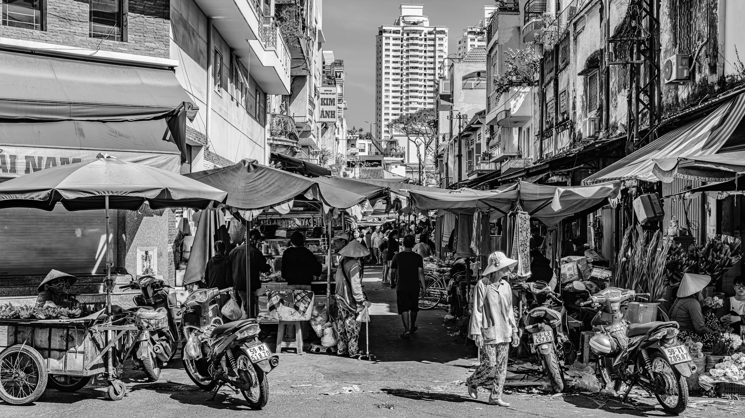 Street market scene with vendors selling goods under tents, motorbikes parked along the street, and people walking. Multi-story buildings line the street in an urban setting.