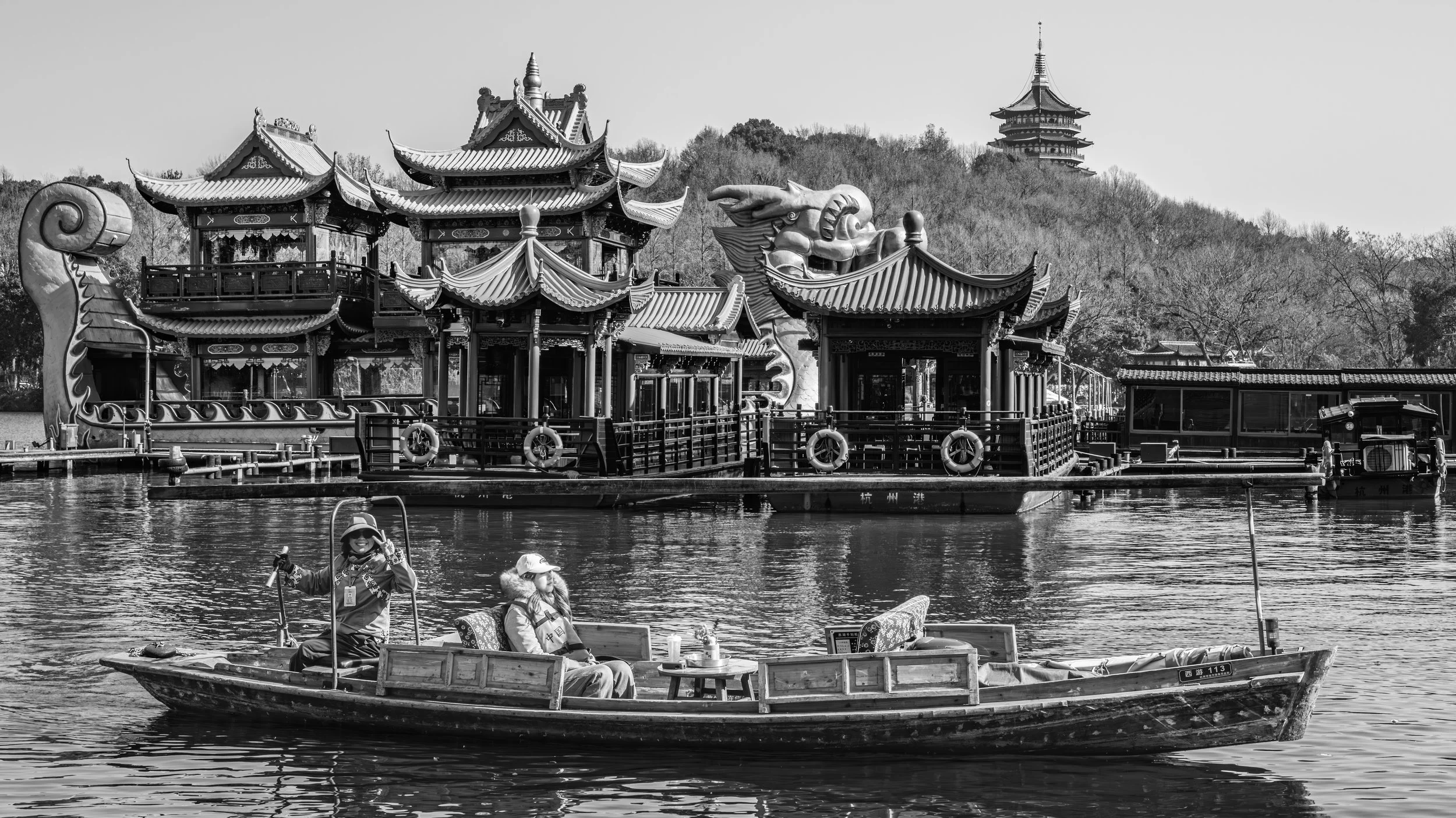 A black and white photo of a traditional Asian-style boat on a lake with people on board, and a large ornate temple structure with multiple rooftops and a dragon sculpture on the boat. In the background. Hangzhou, China.
