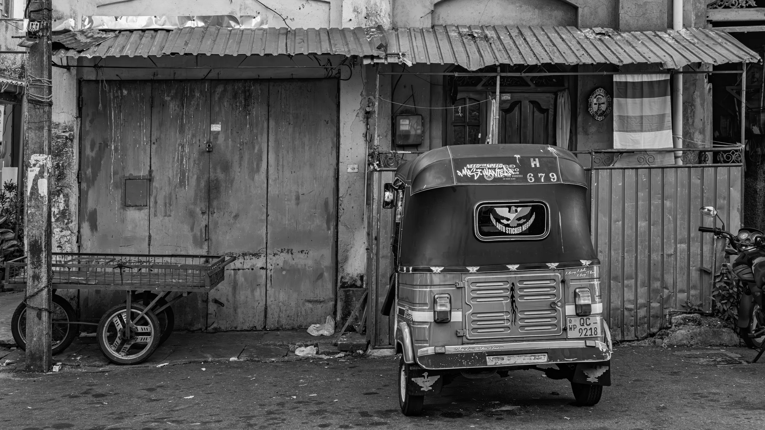 A street scene in black and white showing a small auto rickshaw, a bicycle, and a scooter parked in front of an old building with a corrugated metal roof, a flag, and a small television mounted on the wall.