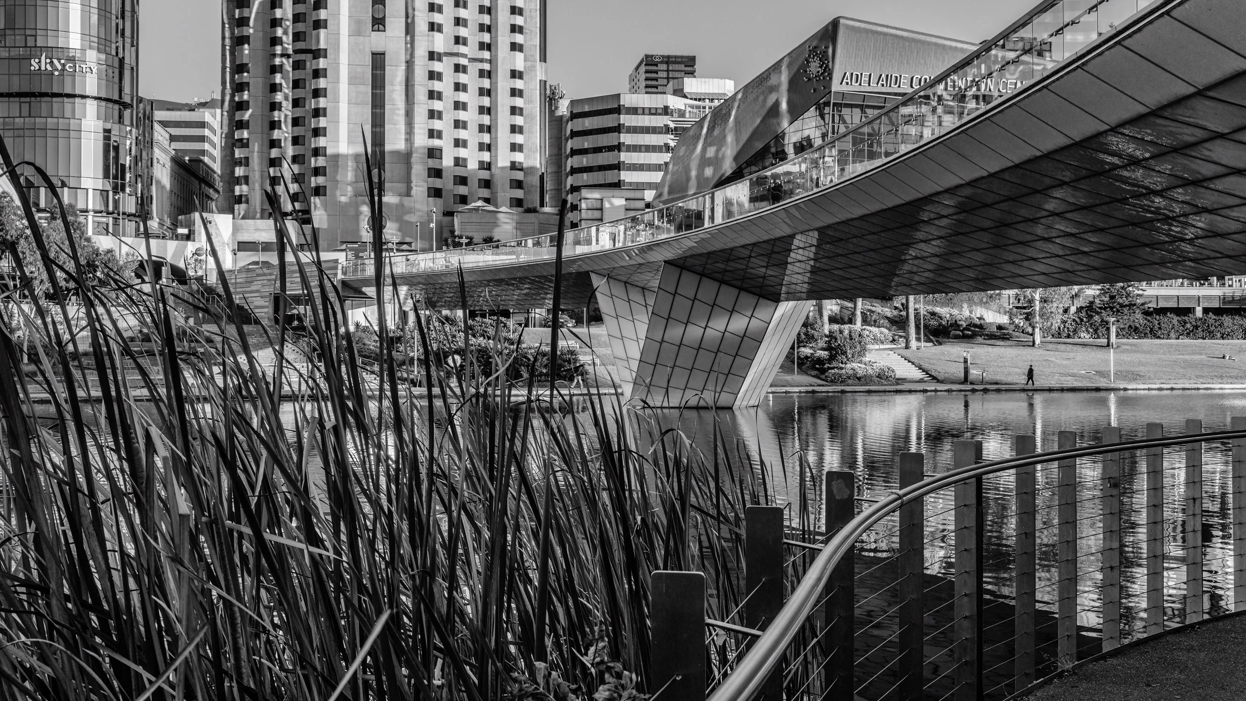 A black and white photo of a modern cityscape featuring tall buildings, a unique contemporary bridge with a reflective surface over a water body, and surrounding greenery.