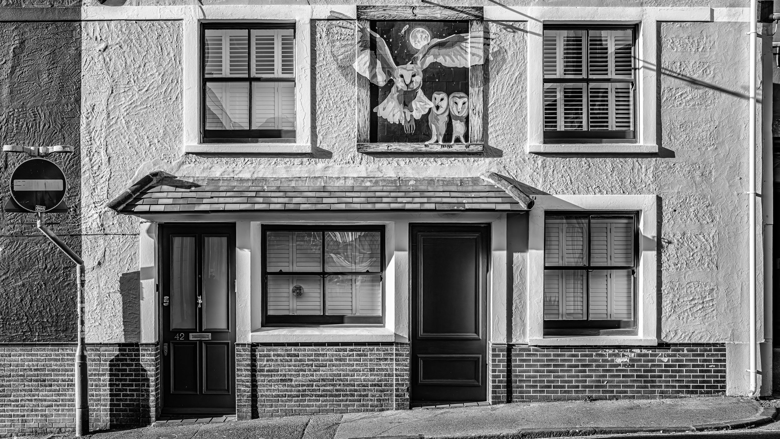 Black and white photo of a two-story residential building with four windows, two on each floor, with shutters on the upper windows. There is a small entrance door at the ground level and a brick lower facade. A street lamp and a no-entry traffic sign