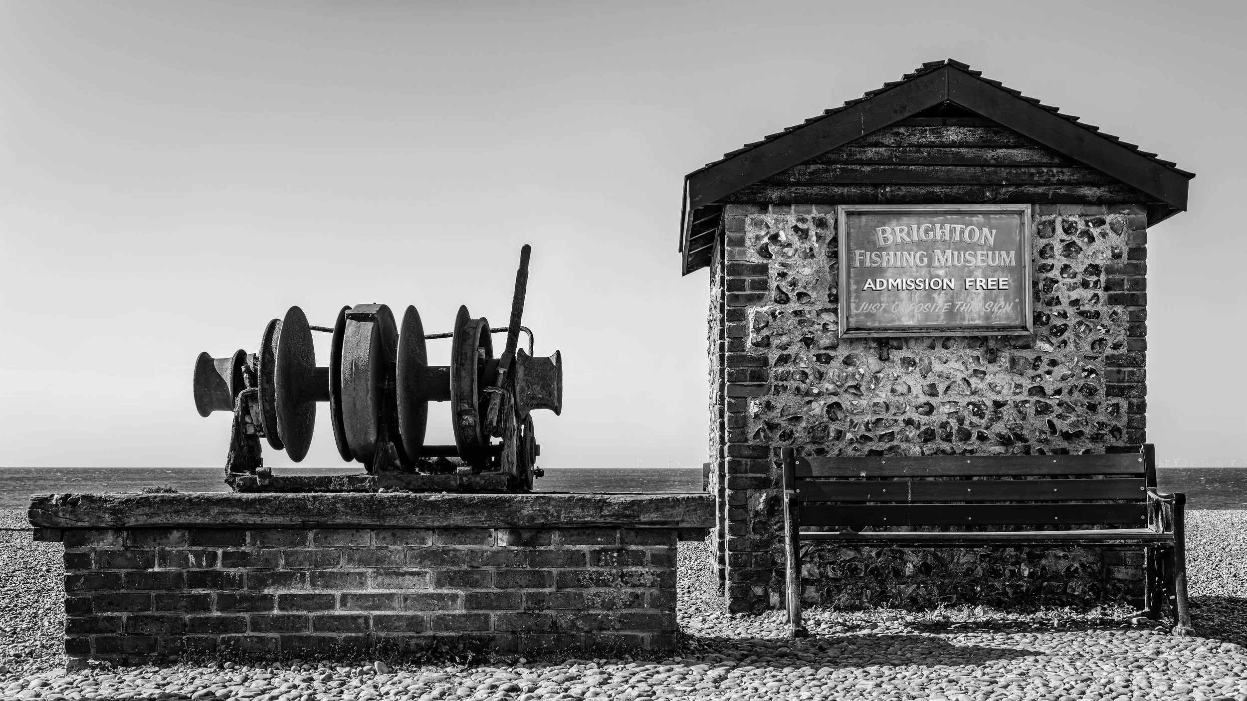 A black and white photo of an old rusted boat winch and a small brick building with a wooden roof on a beach, with a sign for Brighton Fishing Museum that says 'Admission Free'.