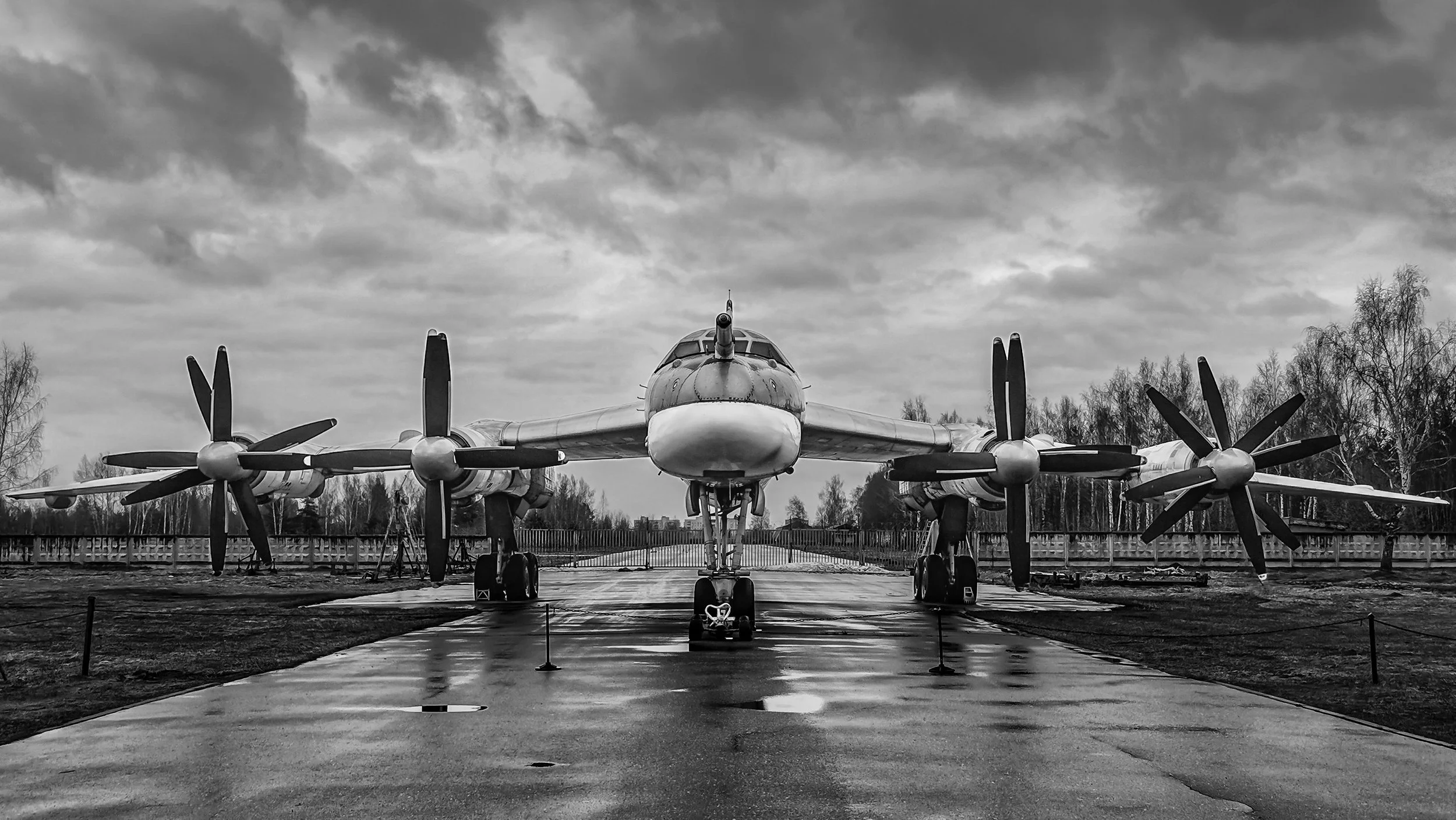 A vintage military aircraft with four propellers on each wing, parked outdoors on a concrete surface under a cloudy sky, surrounded by a fence and trees in the background. Moscow, Russia.