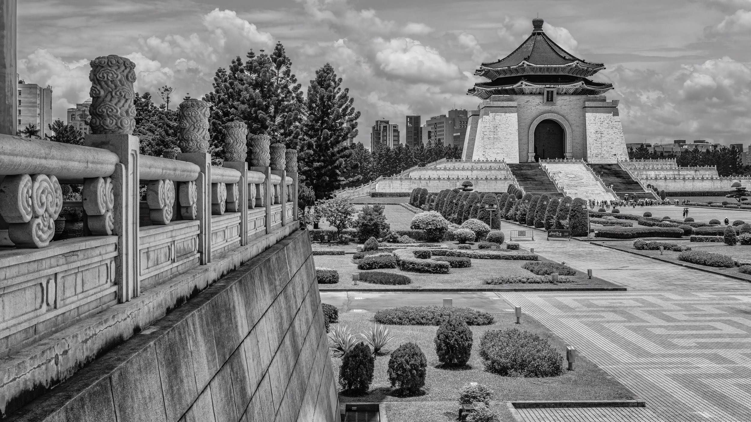 Traditional Chinese architecture with a pagoda-style building on a landscaped garden featuring trimmed bushes, trees, and a stone staircase. The scene is set against a backdrop of modern buildings and a partly cloudy sky. Taipei, Taiwan.