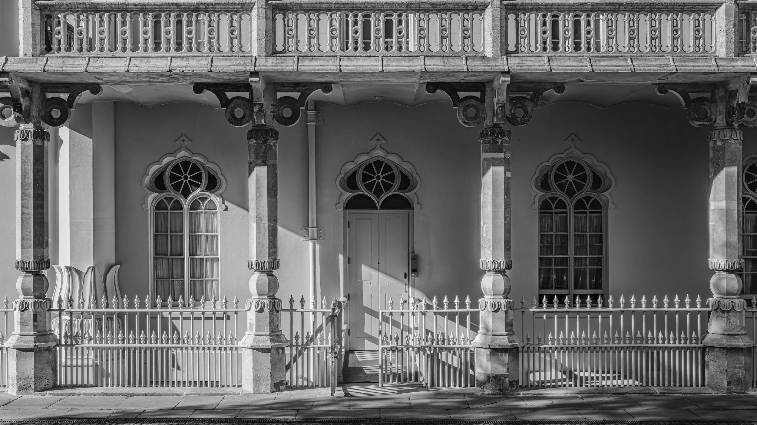 A black-and-white photo of a Victorian-style house with ornate columns, arched windows, and a balcony with a decorative railing.