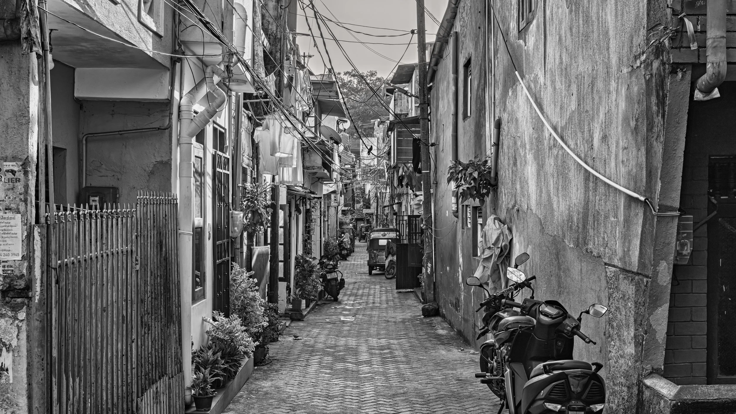 A narrow alleyway in an urban area with residential buildings, motorcycles parked along the side, and numerous electrical wires overhead, in black and white.