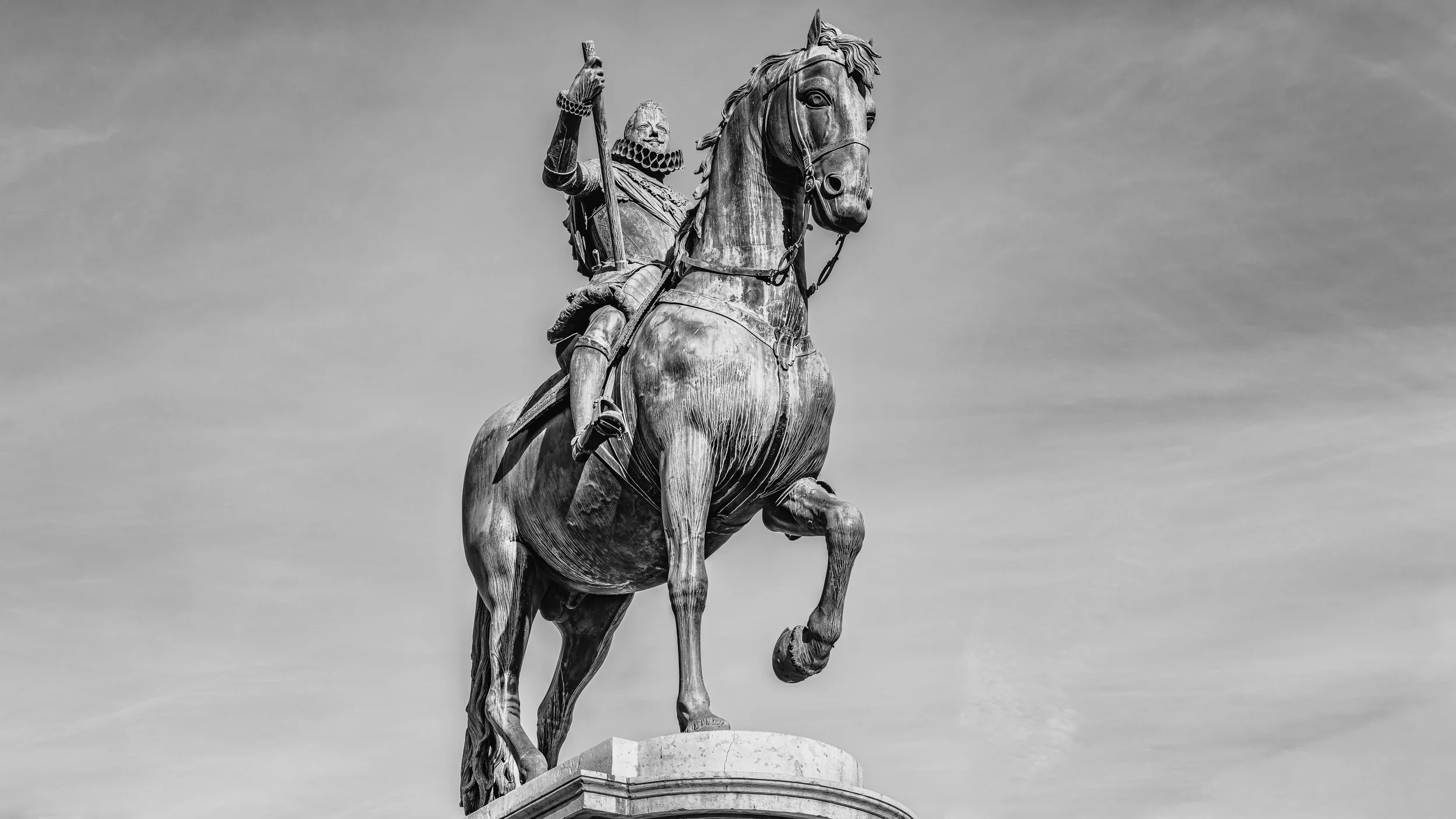 Black and white photo of a large equestrian statue featuring a rider in historical military attire on a horse, set against a cloudy sky.