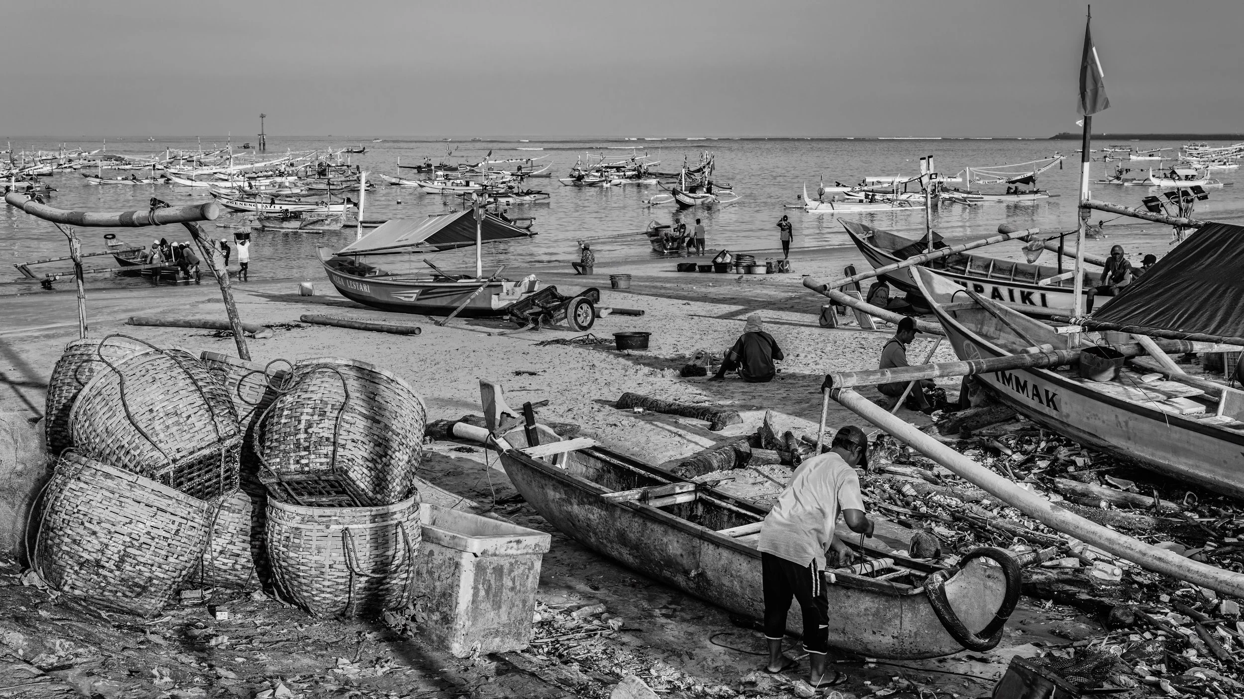 A black and white photo of a busy fishing harbor with numerous boats docked or on the shore, and fishermen working near the water. There are baskets and fishing equipment scattered around.