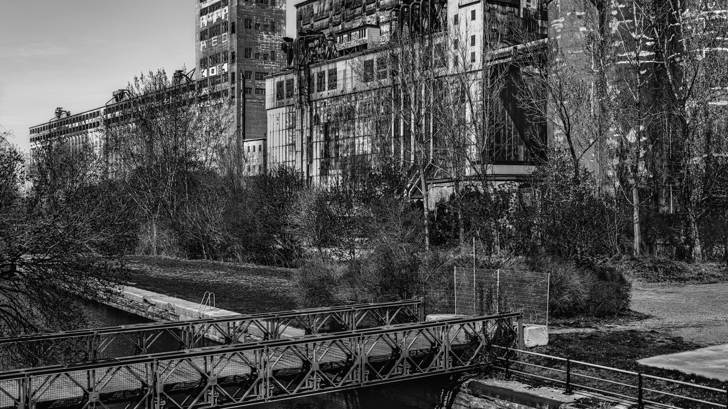 Black and white photo of tall, worn apartment buildings with graffiti, behind leafless trees. A canal with a small bridge crosses in the foreground, with a gravel path and a fence nearby.